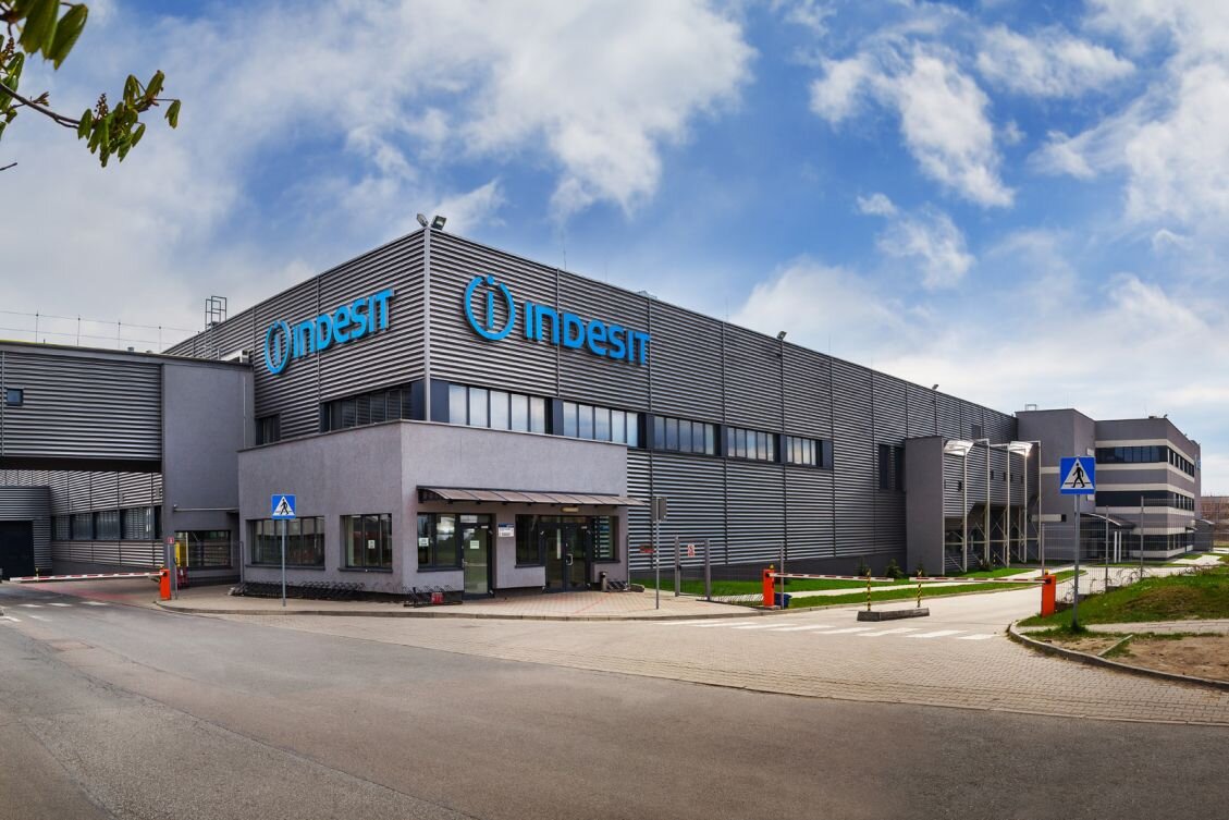 Zdjęcie przedstawia siedzibę firmy Indesit. Gmach firmy to duża, prostokątna bryła. Posiada prostokątne okna ułożone w rzędzie, ściany firmy są zbudowane z szarych paneli. Przed firmą znajduje się wyasfaltowana droga dojazdowa. Wjazd na teren przedsiębiorstwa jest zagrodzony szlabanami.