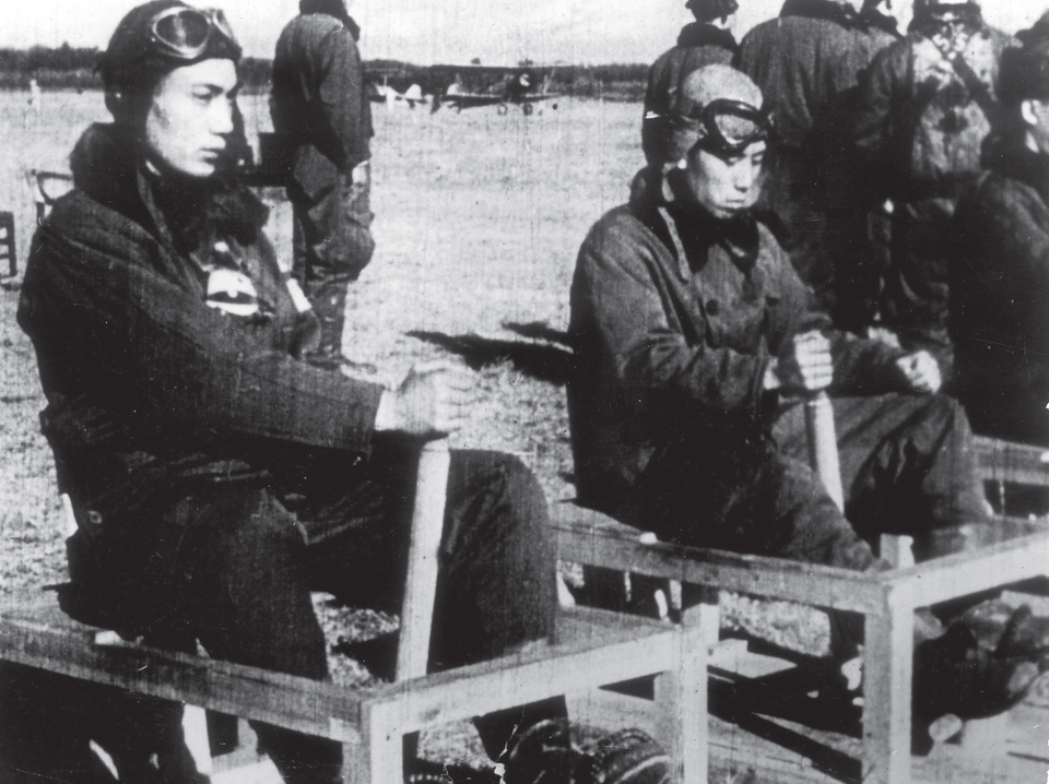 Czarno- białe zdjęcie przedstawi młodych mężczyzn siedzących w drewnianej konstrukcji. Prawą rękę trzymają na wysokim drążku. To japońscy piloci podczas przyspieszonego naziemnego kursu na pilota. 