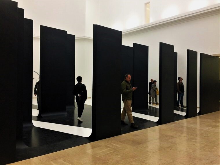 Fotografia przedstawia aranżację przestrzenną w galerii, przypominającą szerokie wstęgi leżące na podłodze, których końce wznoszą się do góry jak wysokie ściany. Pomiędzy nimi przechodzą ludzie.  