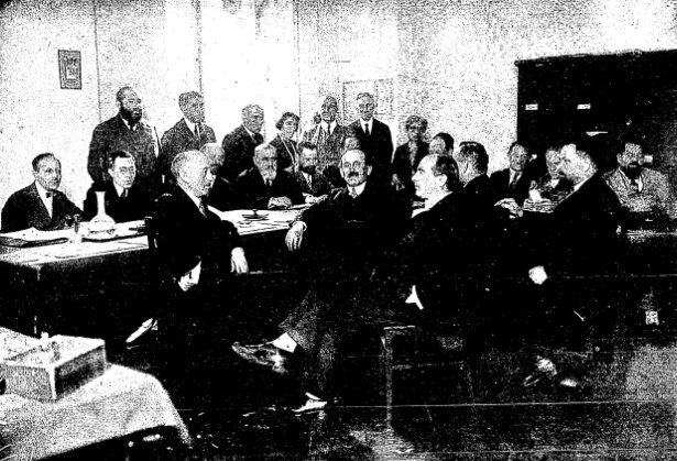 Zdjęcie przedstawia dużą salę, w której znajdują się mężczyźni ubrani w garnitury. Część z nich siedzi, za stołem ustawionym po lewej stronie sali, część na środku sali. Niektórzy stoją pod ścianą za stołem.