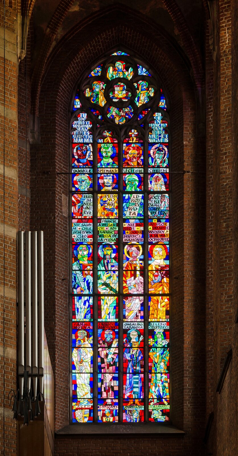 Fotografia przedstawia kolorowy witraż umieszczony na szybie nieopodal piszczałek organowych. Witraż został wykonany z kolorowych kawałków szkła - dominują kolory: czerwony, żółty, niebieski i zielony. Na witrażu przedstawione zostały postaci świętych - osiem pełnych postaci oraz cztery głowy. Pod każdym wizerunkiem umieszczony został tekstowy fragment modlitwy.