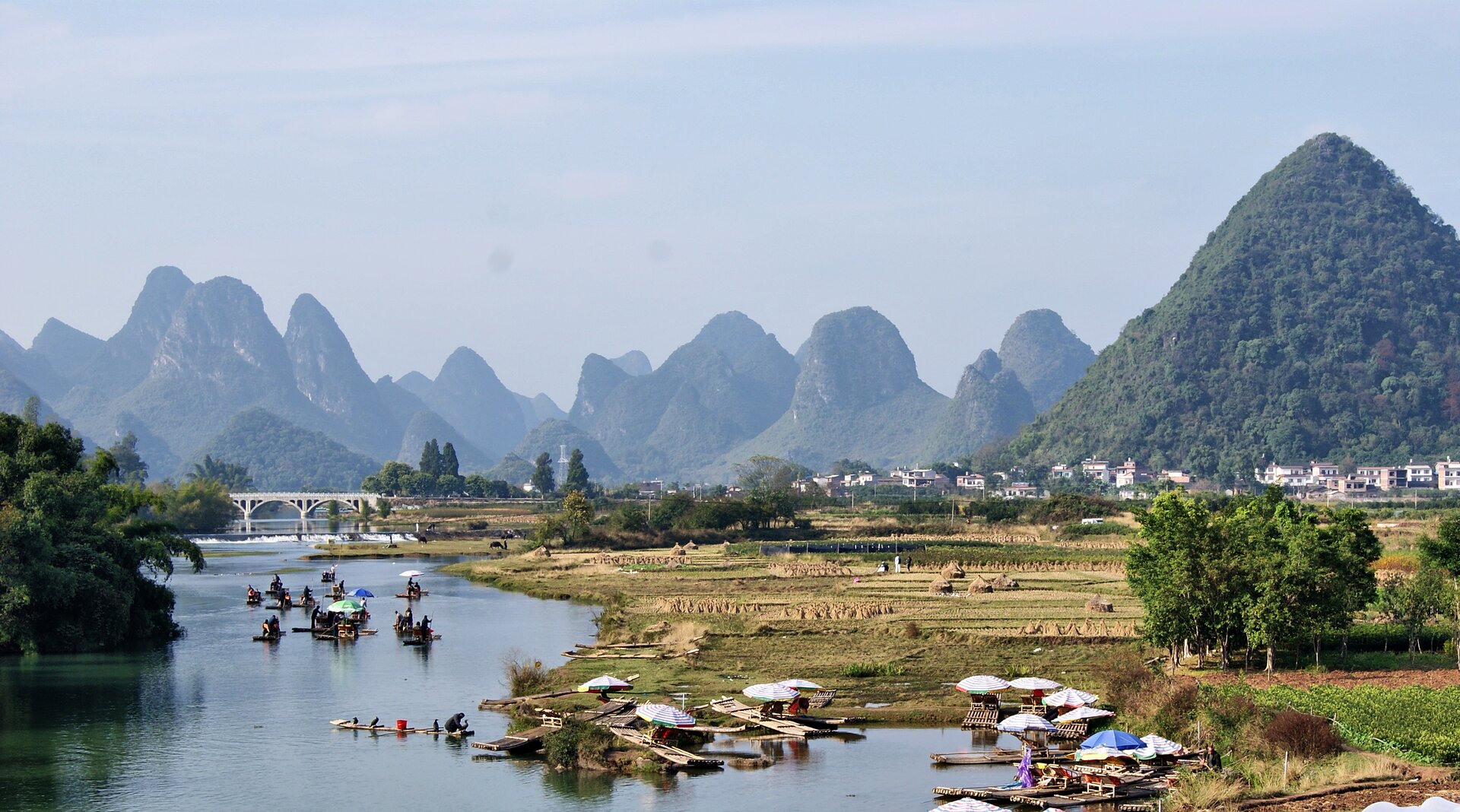 Na zdjęciu znajduje się płynąca rzeka, a na niej dużą ilość małych łódek. Na prawym brzegu widać małe, przycumowane łódki i rozłożone parasole, a w oddali duży, biały most. Po prawej stronie rzeki znajduje się płaski, piaszczysty teren i zabudowania miasta. W tle widać wysokie, stożkowe wzgórza o okrągłych wierzchołkach. Wzgórza pokryte są lasem, nad nimi niebieskie niebo.