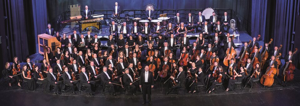 Ilustracja przedstawia fotografię Michała Hellera, prezentującą orkiestrę OiFP w 60. Jubileuszowym Sezonie Artystycznym. Na scenie cała orkiestra. Muzycy siedzą na swoich miejscach. Z przodu instrumenty smyczkowe: skrzypce, altówki, wiolonczele. Za wiolonczelami kontrabasy. Za instrumentami smyczkowymi - instrumenty dęte. Z tyłu, na podwyższeniu instrumenty perkusyjne. Przed orkiestrą przodem stoi dyrygent.