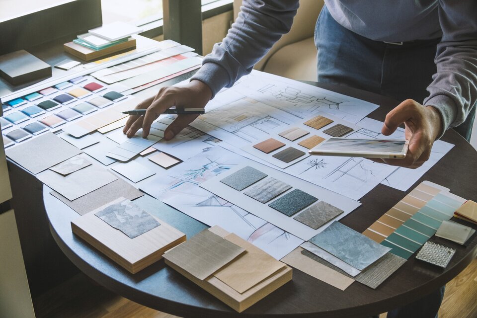 Zdjęcie przedstawia projektanta wnętrz podczas pracy. Mężczyzna ma na sobie niebieskie jeansy oraz jasną, szarą bluzę z długim rękawem. Znajduje się w pomieszczeniu, stoi przy brązowym, drewnianym, okrągłym stole, na którym znajduje się mnóstwo próbek materiałów. Projektant w lewej dłoni trzyma tablet, a w prawej długopis.