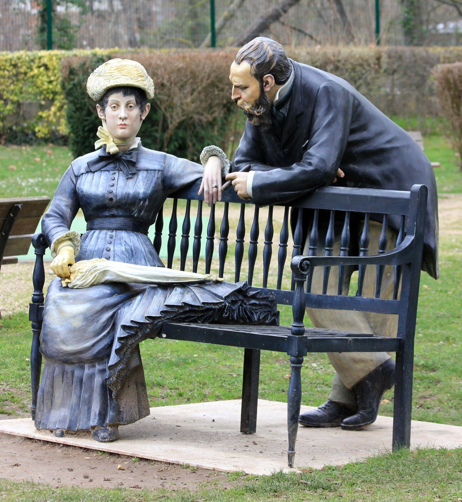 Zdjęcie przedstawia dwie rzeźby – jedną przedstawiającą siedzącą kobietę na ławce, a drugą mężczyznę opartego o tę ławkę. Mężczyzna ma skierowane oblicze w stronę kobiety, która wyraźnie nie zwraca na niego uwagi.