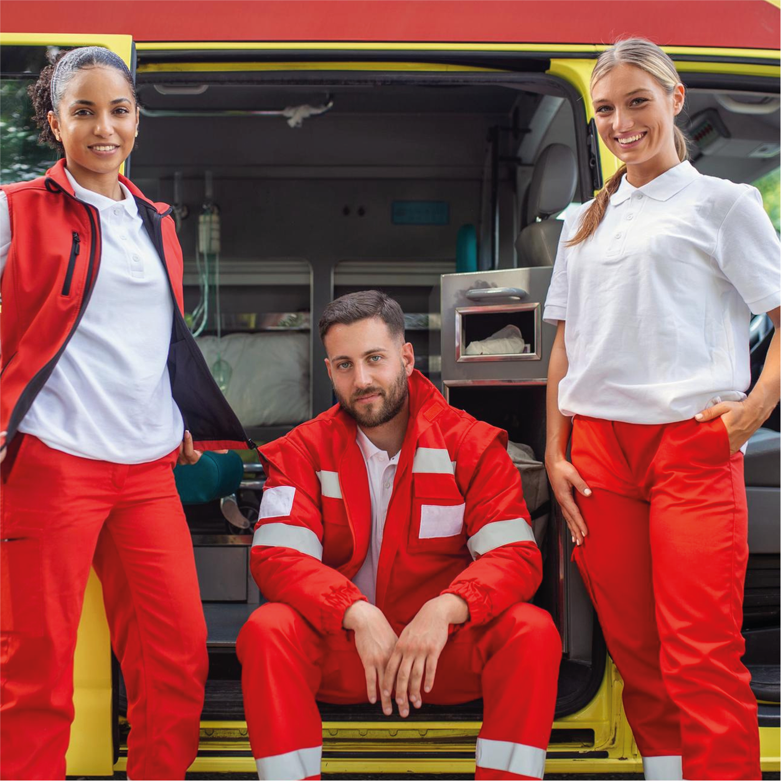 Zdjęcie przedstawia troje ratowników medycznych w biało‑czerwonych strojach, pozujących na tle karetki. Pomiędzy dwiema stojącymi kobietami siedzi mężczyzna. 