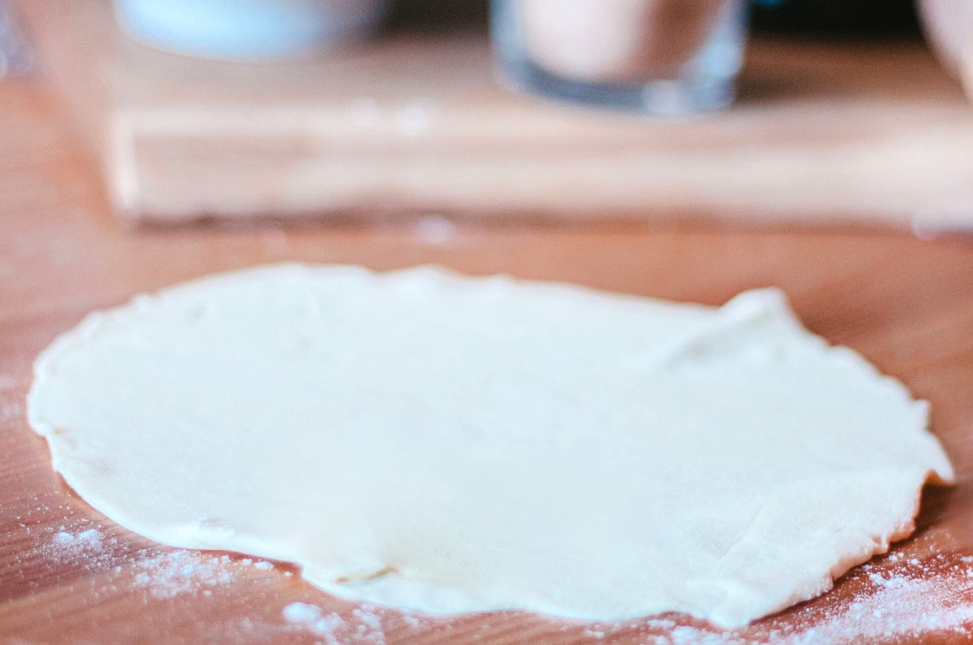 Zdjęcie przedstawia krok drugi formowania wianka debrzeńskiego. Zdjęcie wykonane jest od góry pod skosem. Widoczny jest jasny okrągły rozwałkowany placek ciasta, leżący na drewnianym ciemnobrązowym blacie. Dookoła ciasta rozsypana jest biała mąka. W tle dostrzec można niewyraźny zarys drewnianej jasnobrązowej deski na której stoi jasny pojemnik oraz szklanka z jajkiem w środku.