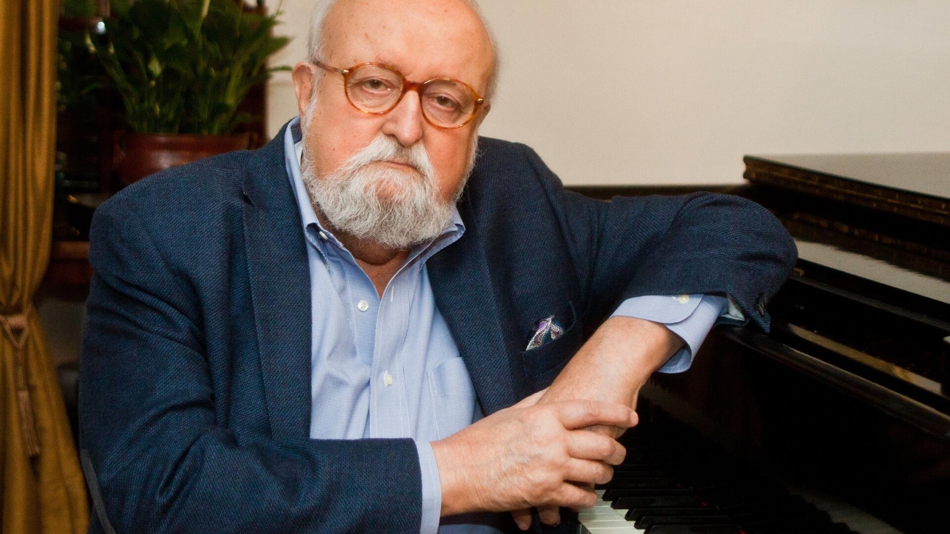 Ilustracja przedstawia fotografię Krzysztofa Pendereckiego. Mężczyzna opiera się o czarny fortepian. Ubrany jest w koszulę oraz marynarkę.