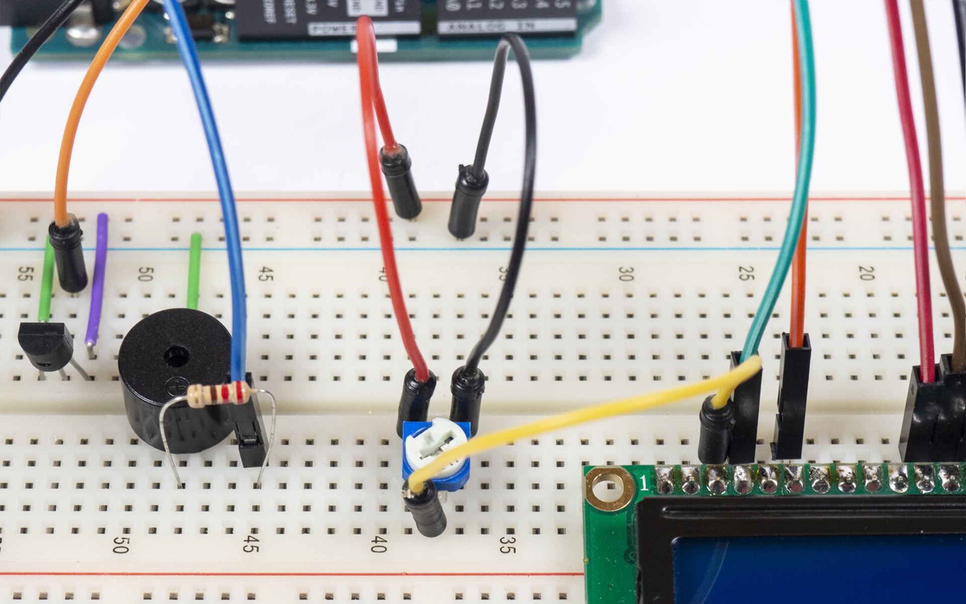 Zdjęcie przedstawia połączenia pomiędzy arduino uno, a płytką stykową. Na płytce znajduje się termometr (względem wycięcia w obudowie) podłączony jest prawą nóżką do uziemienia, lewą do +5V a środkową do pinu A0 arduino. Pin 5V podłączony jest do +5V płytki, a GND podłączone jest do uziemienia. Uziemienie górnej szyny jak i dolnej na płytce stykowej jest połączone. Podłączona jest także LED, jedną nóżką do uziemienia, drugą zaś poprzez rezystor do pinu 7 arduino. Po środku płytki zamontowany jest brzęczyk, który poprzez rezystor łączy się z pinem 6 arduino, a drugą nóżką do uziemienia. Po prawej stronie płytki stykowej podłączono ekran LCD, do którego podłączono kable prowadzące do arduino. Pomiędzy ekranem a brzęczykiem zamontowano potencjometr, do jego nóżek podłączono kable, środkowy do 3 pinu ekranu, lewa nóżka do +5V natomiast prawy do uziemienia.