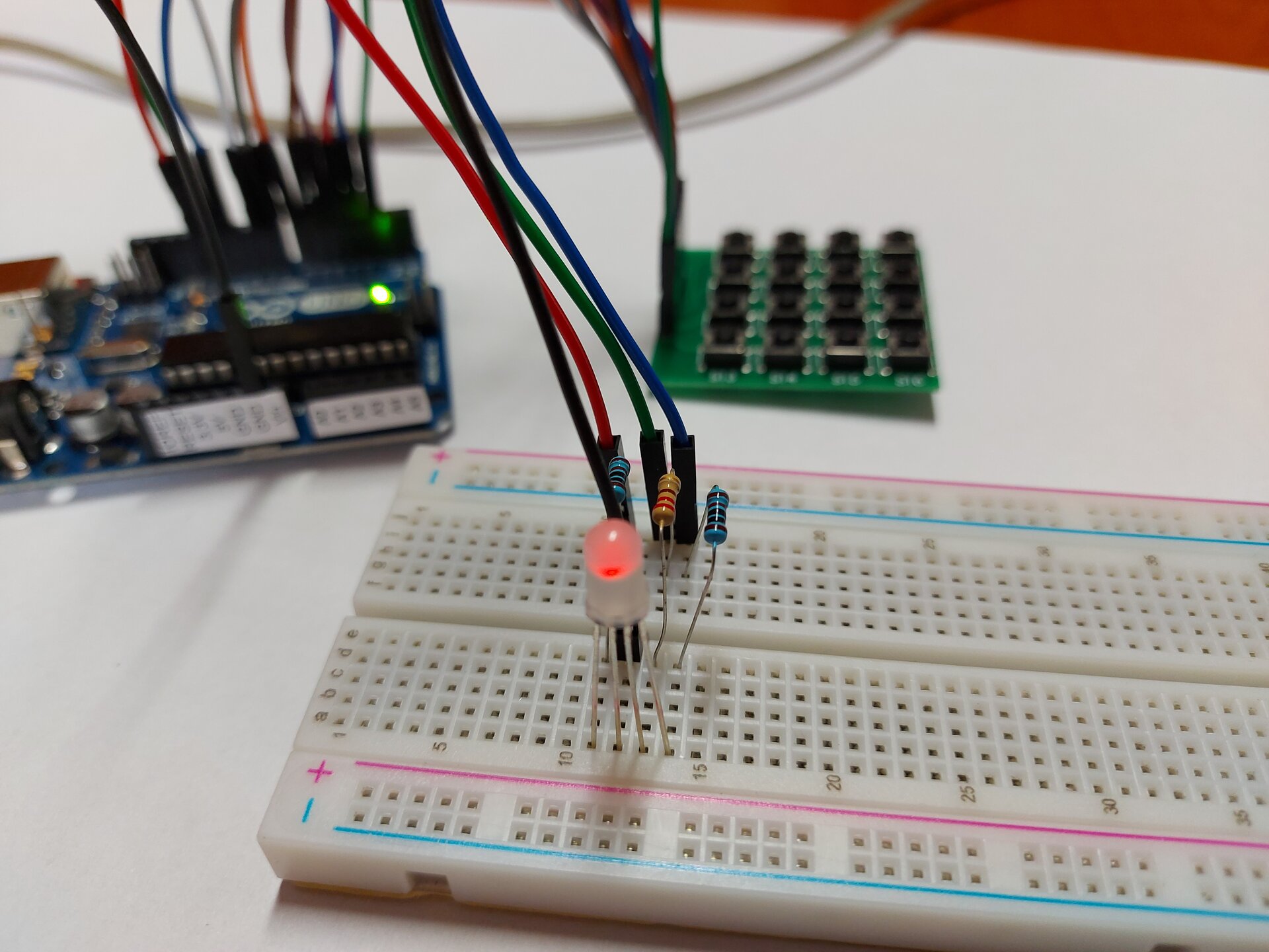 Zdjęcie przedstawia arduino uno połączone kablami do płytki stykowej, na której znajdują się 3 rezystory podłączone do kolejnych nóżek 4 pinowej diody, ostatnia nóżka podłączona jest kablem do arduino. W tle widoczna jest płytka z przyciskami, która podłączona jest do arduino osobnym zestawem kabli. LED świeci się na czerwono.
