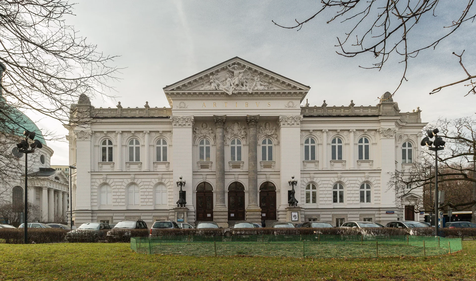 Na zdjęciu biały potężny budynek o bogatych ornamentach. Wysuniętą do przodu część wspierają dwie wielkie kolumny, na których spoczywa tympanon. Przy wejściu do obiektu zarówno po lewej, jak i prawej stronie znajdują się zdobne lampy. Przed gmachem rośnie zadbana trawa.