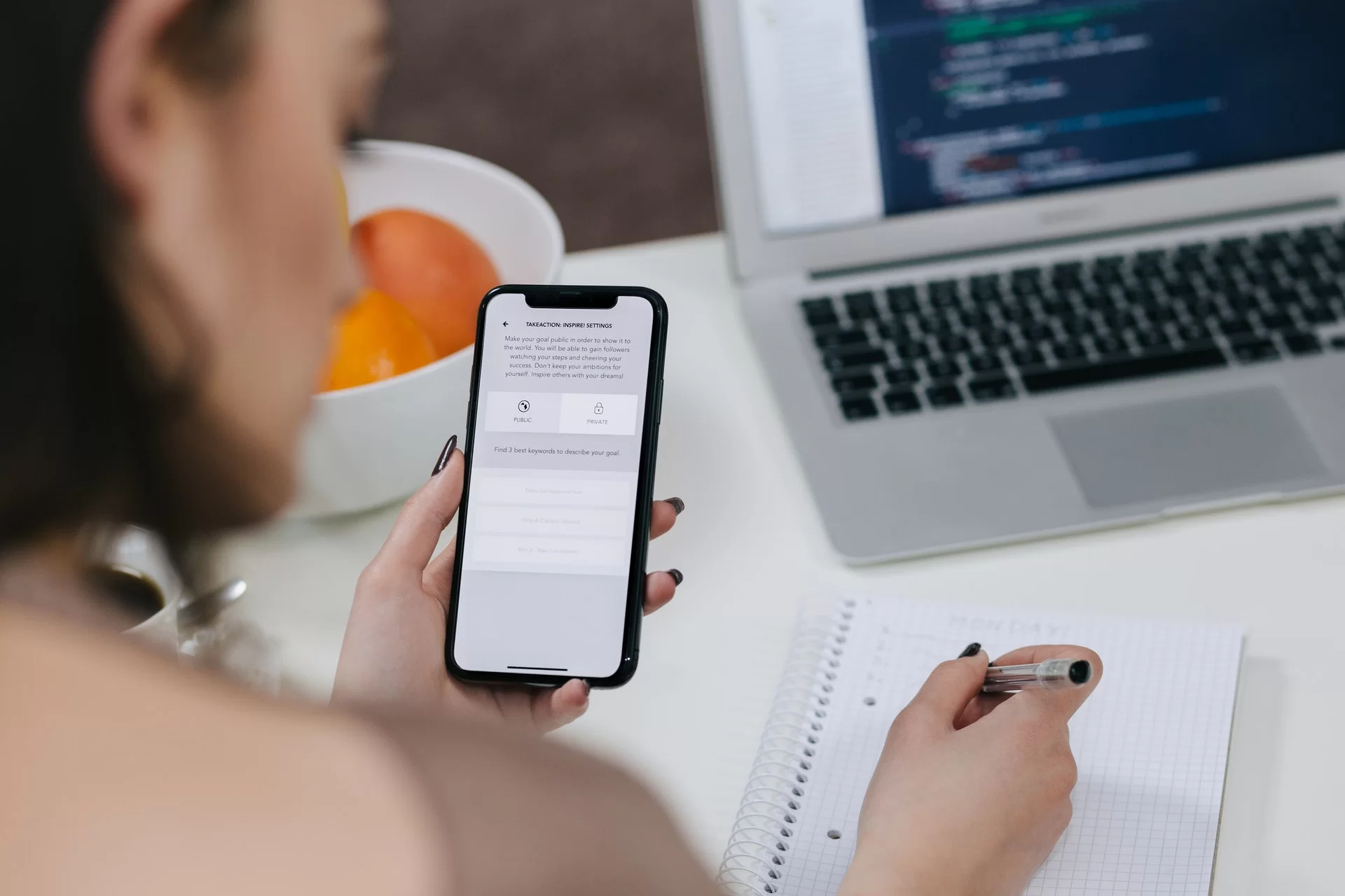Zdjęcie przedstawia kobietę, która siedzi przy biurku. W lewej ręce trzyma włączony smartfon, a prawą ręką pisze długopisem w zeszycie. Na biurku stoi laptop oraz miska z owocami.