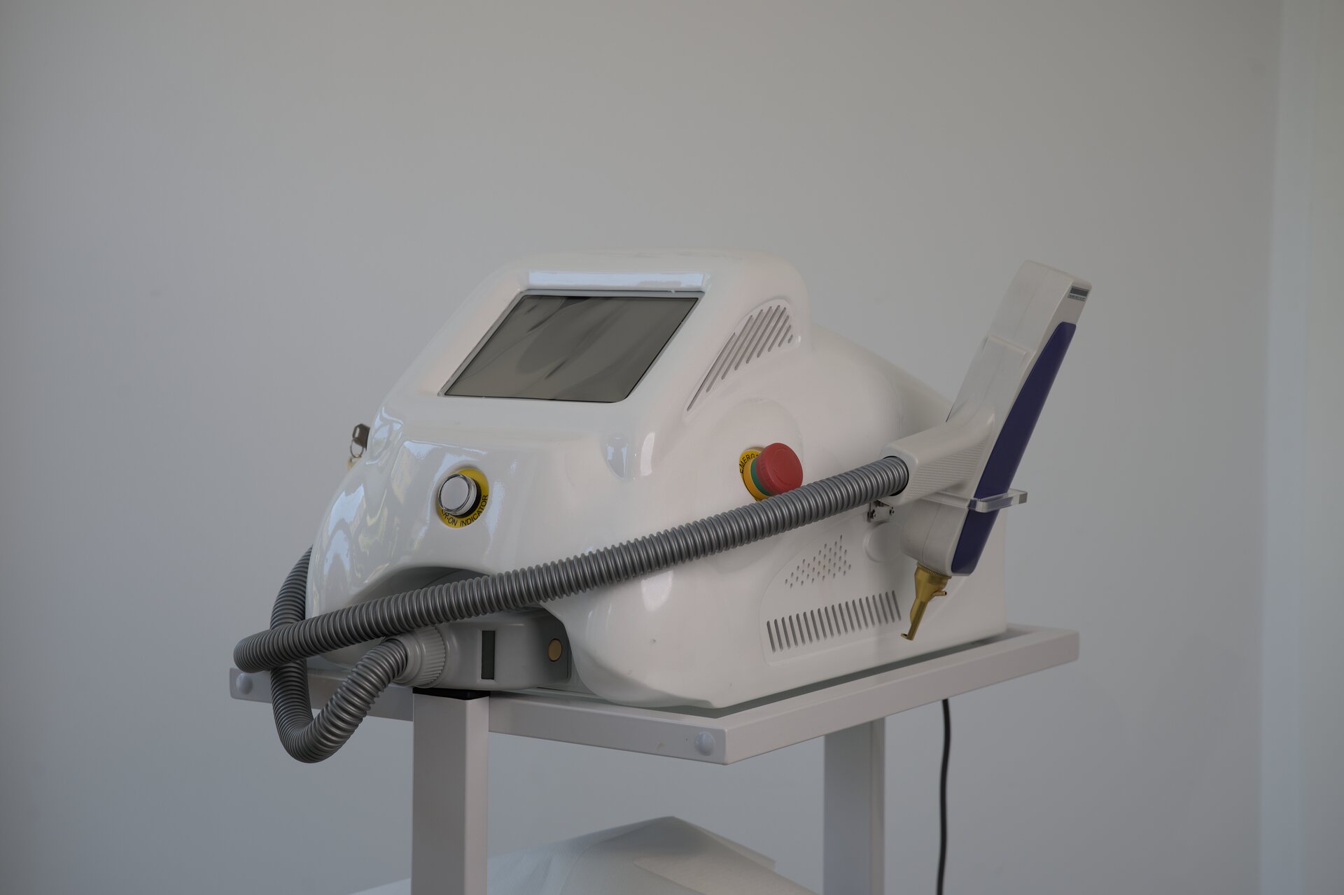 Zdjęcie przedstawia laser. Urządzenie składa się z korpusu w białej obudowie z dużym ekranem oraz dwoma przyciskami. Za pomocą grubego przewodu do korpusu podłączona jest głowica lasera, która wisi na wieszaku z boku urządzenia. Głowica lasera składa się z rączki oraz metalowej, wąskiej końcówki.