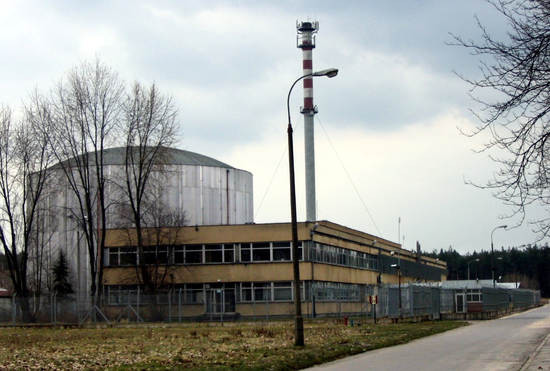 Zdjęcie przedstawia szary budynek o cylindrycznym kształcie oraz drugi niższy, prostokątny, koloru żółtego. W tle znajduje się biało‑czerwony maszt radiowy.  