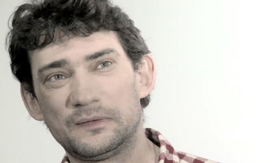 Zdjęcie przedstawia mężczyznę z ciemnymi, lekko kręconymi włosami, krótkim zarostem na kwadratowej twarzy oraz koszulą w biało‑czerwoną kratkę. Tło zdjęcia jest białe.