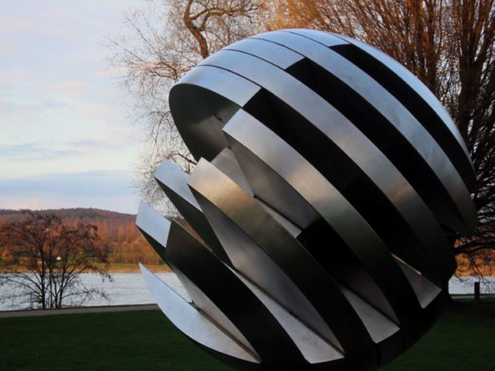 Zdjęcie przedstawia rzeźbę nowoczesną w naturalnej przestrzeni nad rzeką Ren, w Niemczech. Rzeźba w całości wykonana jest z polerowanego metalu, w formie ażurowej kuli. Kula -  dużych rozmiarów jest ponacinana w równoległe plastry,  z których tylko część tworzy formę, a wycięte fragmenty pozostawiły wolne przestrzenie.