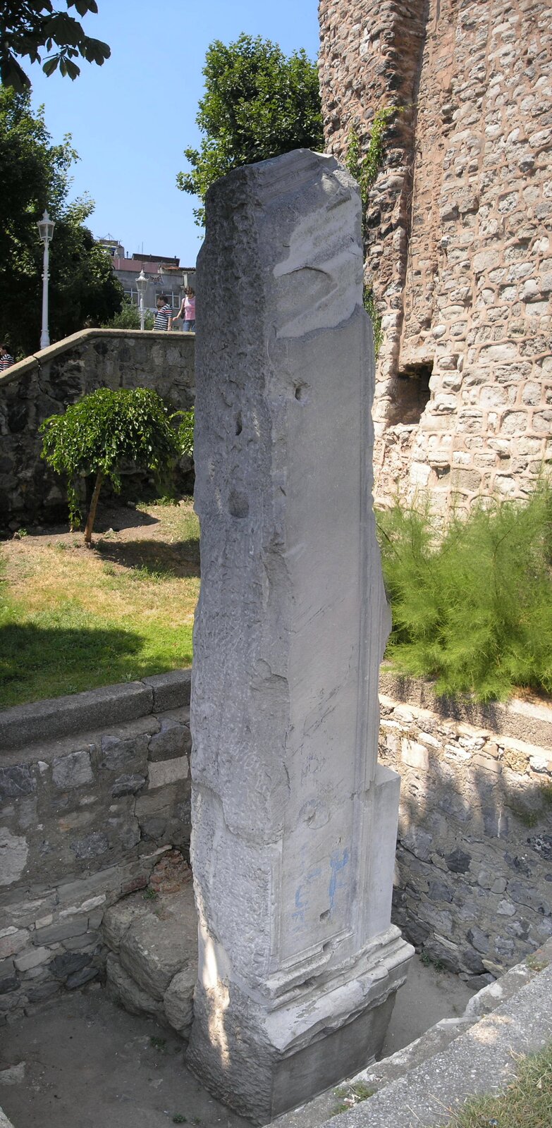 Zdjęcie przedstawia mocno zniszczoną kolumnę stojącą w pobliżu starego muru wykonanego z kamieni. Kolumna jest wysoka, o kształcie prostopadłościanu.