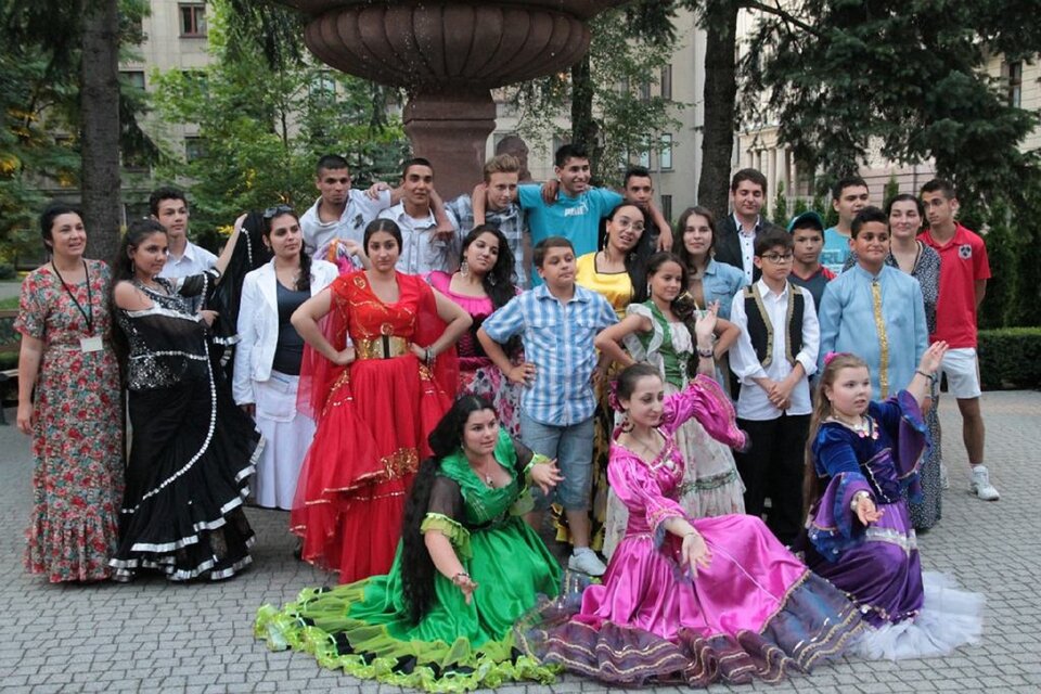 Ilustracja przedstawia grupę żeńską Romów. Część z nich ubrana jest w charakterystyczne stroje cygańskie. Kobiety mają długie, kolorowe suknie z falbanami. W tle widoczne są drzewa oraz fontanna.