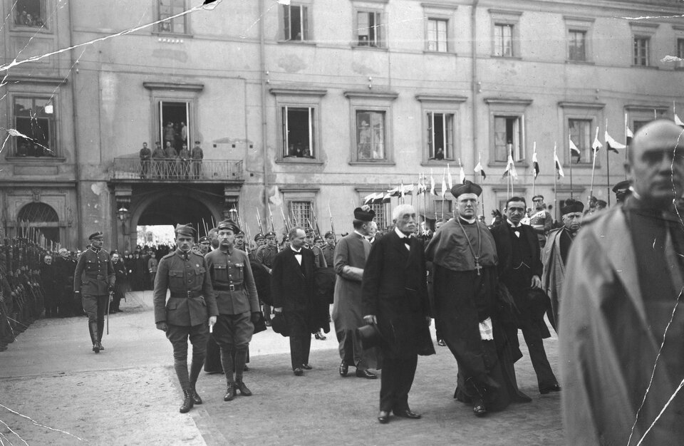 Czarno‑białe zdjęcie przedstawia liczne osoby idące w pobliżu budynku. To mężczyźni w mundurach, duchowni. W tle w rzędzie stoją żołnierze, widać powiewające polskie flagi.  