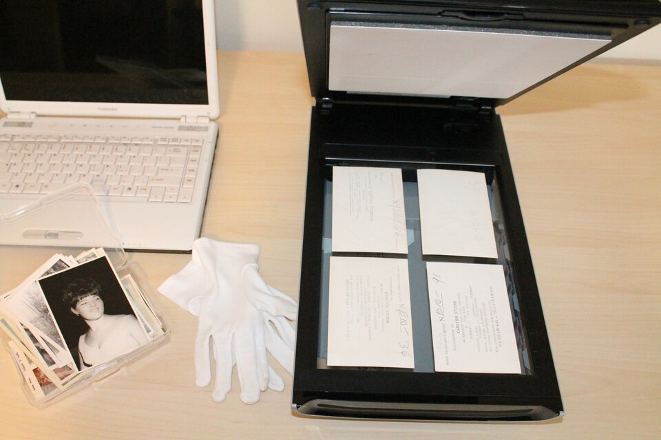 Zdjęcie przedstawia otwarty laptop, obok którego znajduje się skaner z otwartą pokrywą. Leżą na nim cztery prostokątne zdjęcia przygotowane do skanowania. Obok skanera leży para białych rękawiczek i plik starych zdjęć w plastikowej torebce.