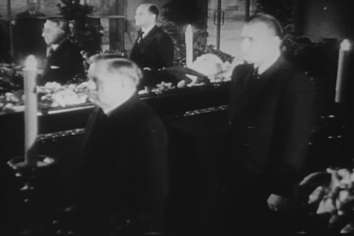 Zdjęcie przedstawia czuwanie przy trumnie Jana Masaryka. Widać otwartą trumnę, w której złożone jest ciało Masaryka. W rogach trumny ustawione są gromnice. Po bokach trumny stoją na baczność czterej mężczyźni w garniturach.