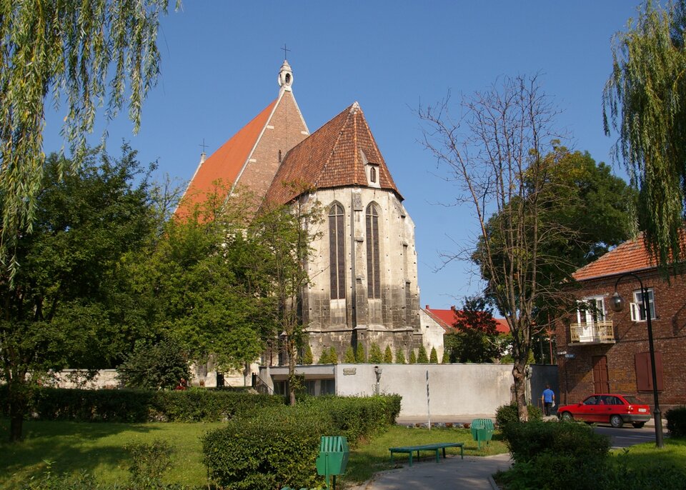 Fotografia przedstawia budynek zbudowany z dwóch brył, obie mają spadziste dachy. Na wyższym budynku widać krzyż. Wokół rosną drzewa, widać zaparkowany samochód i idącego mężczyznę.