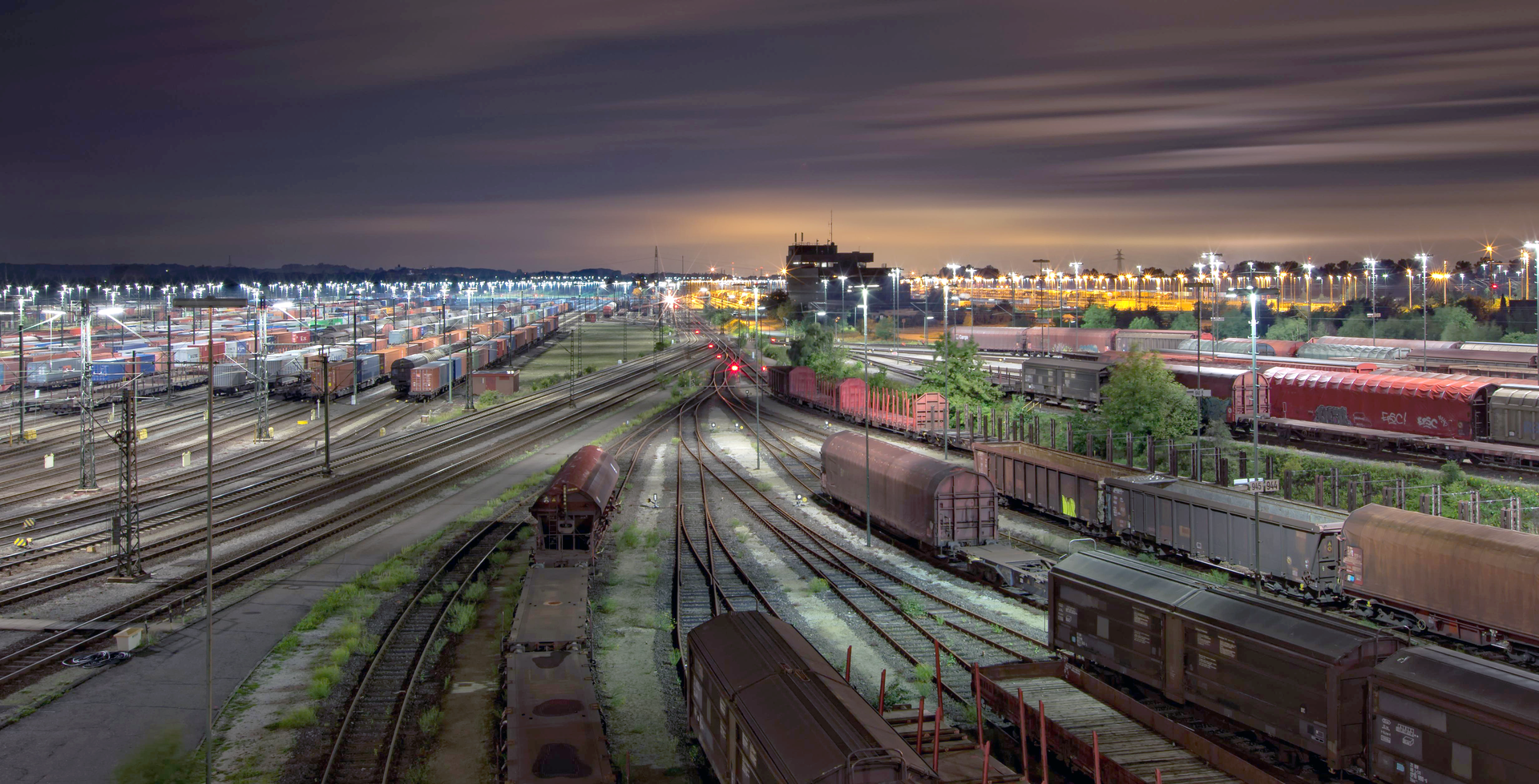 Zdjęcie przedstawia zajezdnię kolejową widoczną wieczorem. Wagony towarowe stoją na torach wszędzie w zasięgu wzroku. Zajezdnia jest dobrze oświetlona lampami na słupach.