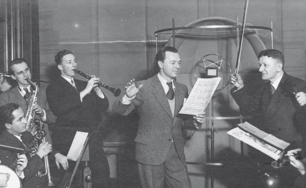 Ilustracja przedstawia członków orkiestry, którzy akompaniują śpiewającemu mężczyźnie. Po lewej siedzi osoba z trąbką, obok niej gitarzysta, dalej saksofonista i klarnecista. Po prawej z uniesionym do góry smyczkiem stoi dyrygent. Centralną postacią jest piosenkarz stojący przed mikrofonem i trzymający w ręku kartki z nutami. Wszyscy zebrani to młodzi mężczyźni ubrani w marynarki, koszule i krawaty.