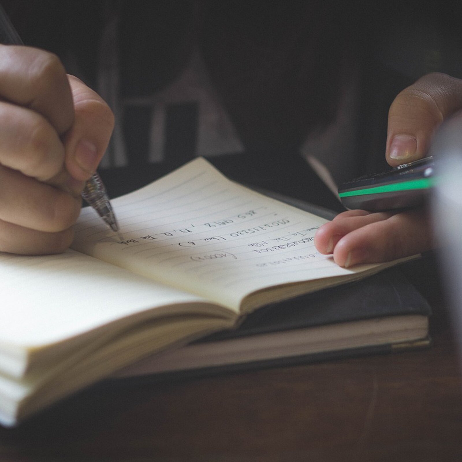 Na zdjęciu widoczne są czyjeś dłonie. Prawą ręką ktoś zapisuje w coś notesie a w lewej klika coś na telefonie komórkowym.