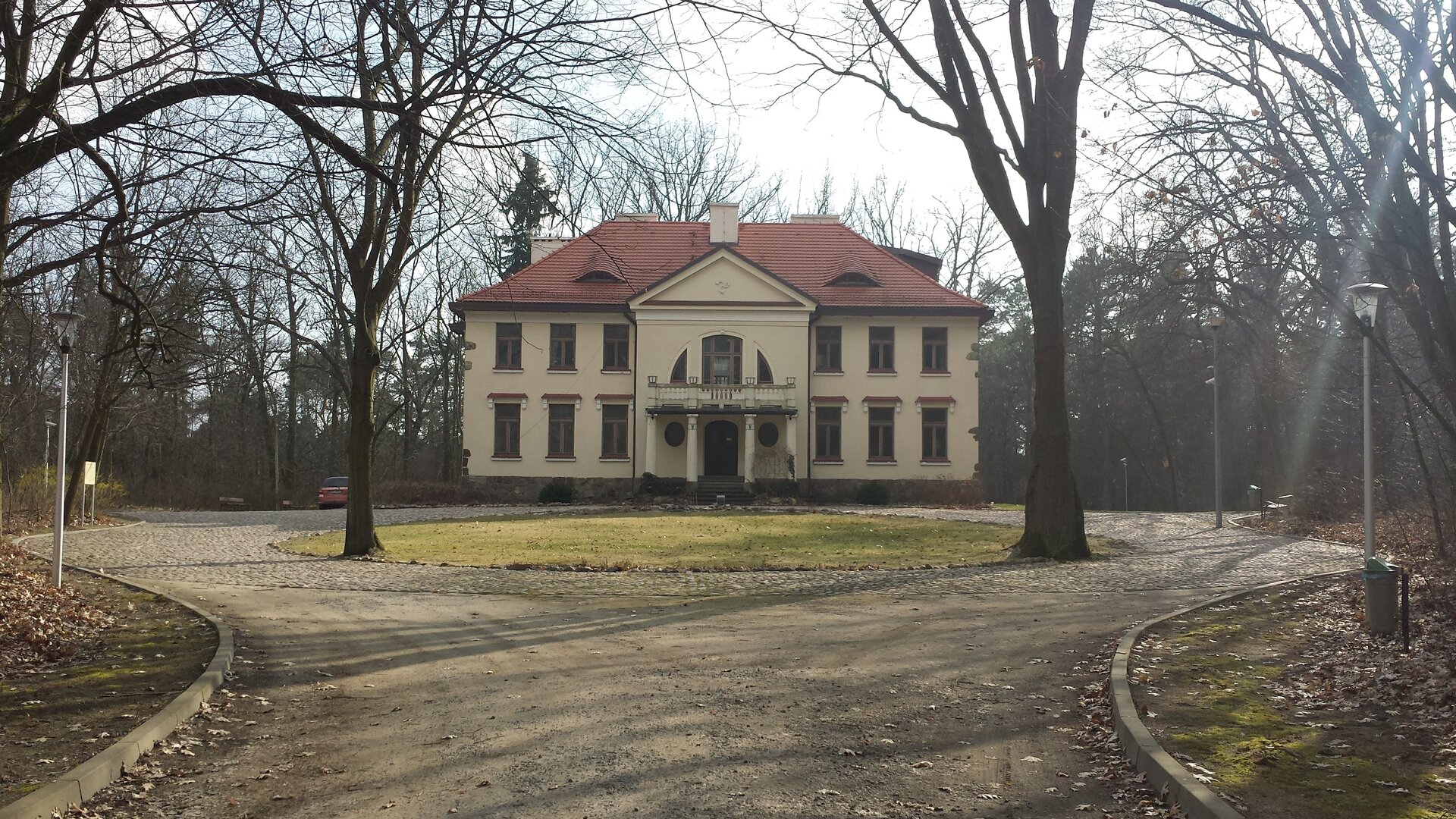 Zdjęcie przedstawia szeroką aleję prowadzącą do dużej, piętrowej willi. Aleja i willa otoczone są parkiem.