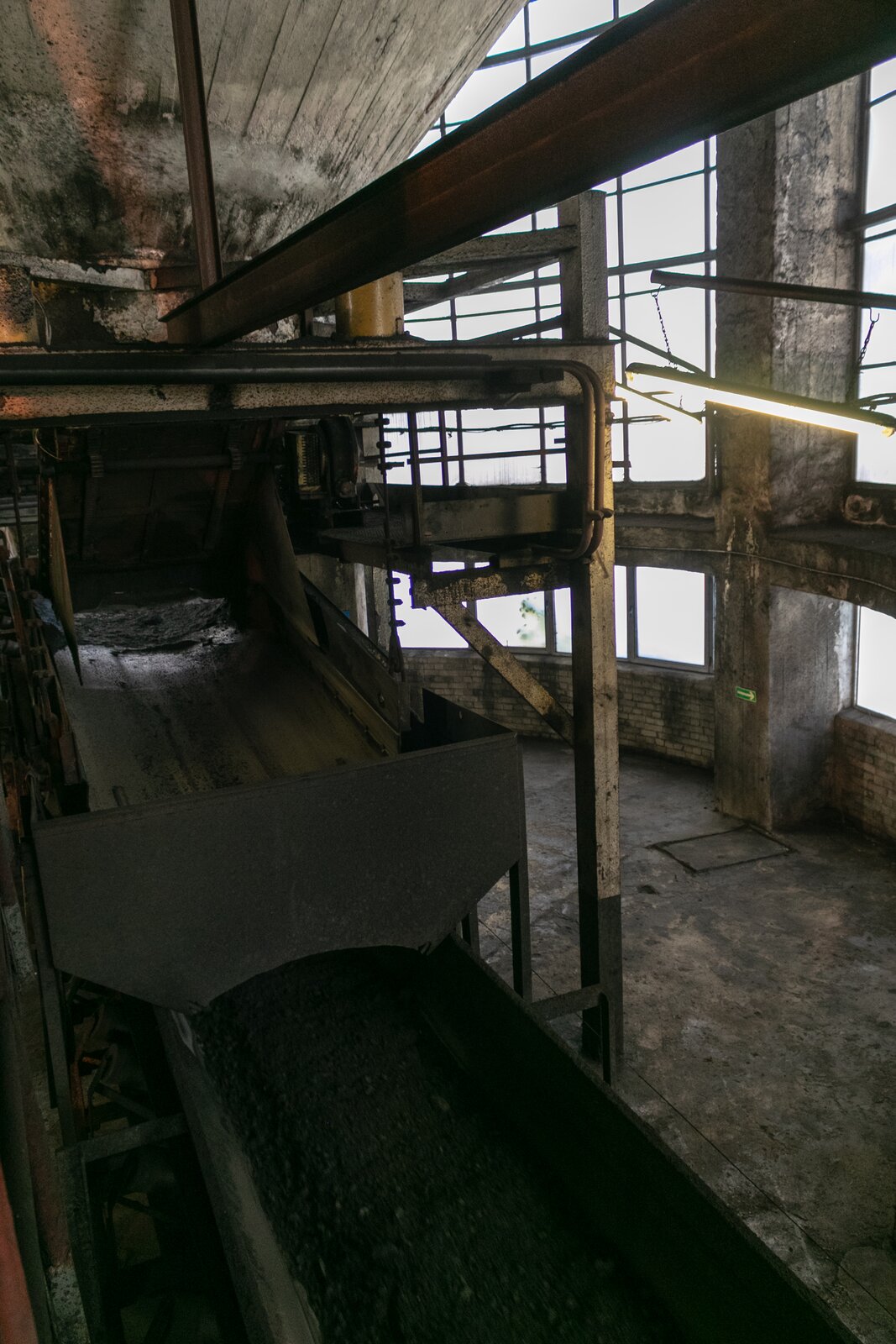 Na zdjęciu widoczne są dwa przenośniki Przenośniki widoczne na zdjęciu są układami modułowym. Na stalowej prostopadłościennej ramię osadzone są lekko nachylonych rolki. Rolki mają kształt walca. Na rolkach osadzona jest taśma. Na taśmie przenośnika widocznego po prawej stronie znajduje się pokruszony węgiel. Boki taśmy są zabezpieczone kratą oczkową uniemożliwiającej wypadnięcie urobku z taśmy. Na górze zdjęcia widać końcówkę taśmociągu. Taśma wchodzi pod gumowe zwisające paski. Oba przenośniki znajdują się pod kątem. Widoczne urządzenia znajdują się w budynku przemysłowym.Poniżej zdjęcia widoczny jest przycisk multimedialny z nagraniem audio. Treść nagrania jest zgodna z tytułem zdjęcia.