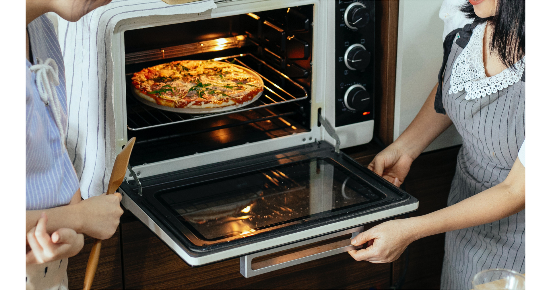 Zdjęcie przedstawia okrągłą pizzę w piekarniku. Obok piekarnika stoją dwie osoby. Jedna z nich otwiera piekarnik.