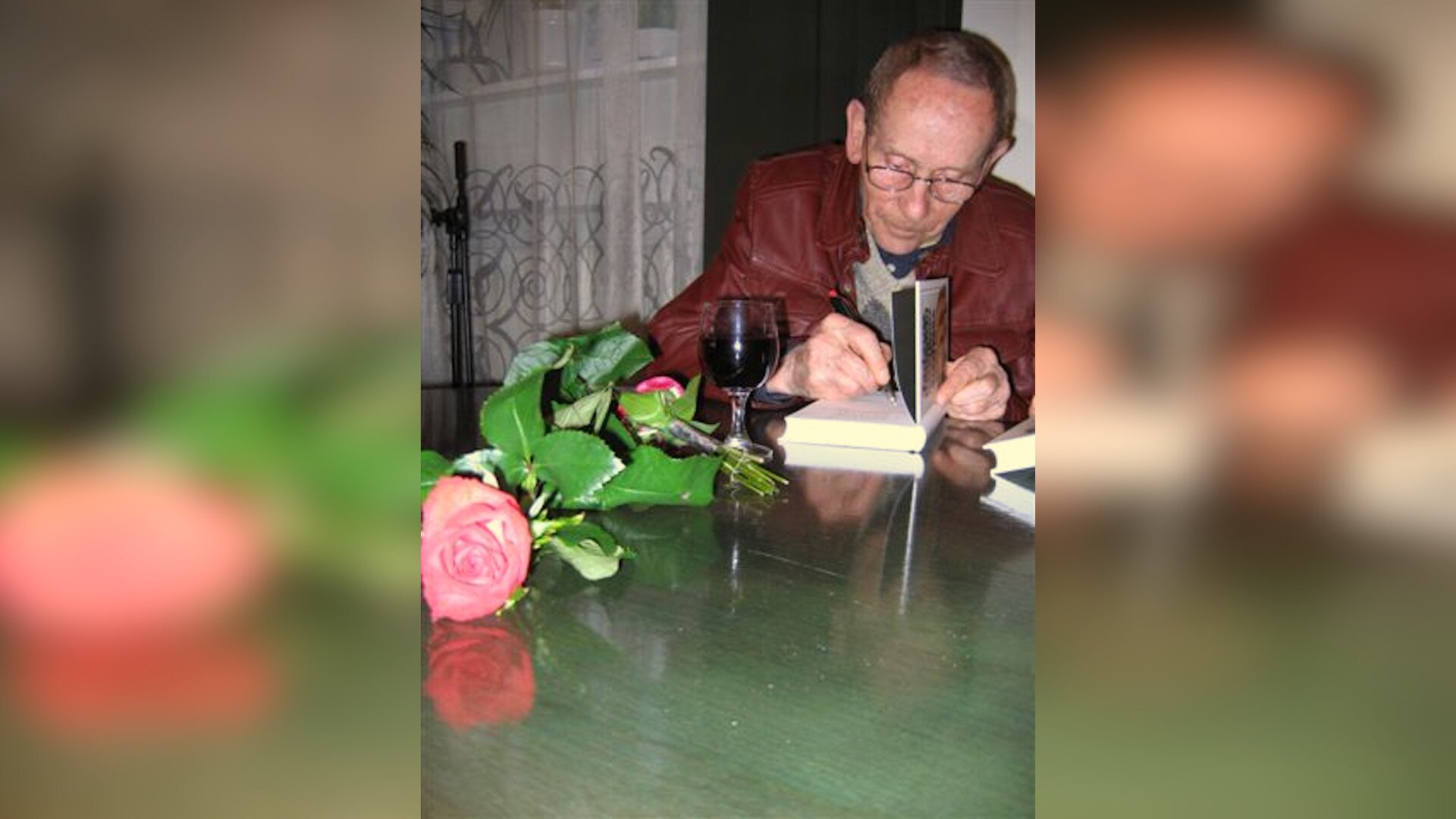 Zdjęcie przedstawia starszego, szczupłego mężczyznę w okularach. Mężczyzna siedzi przy stole i podpisuje książkę. Na stole leży róża i stoi kieliszek czerwonego wina. 