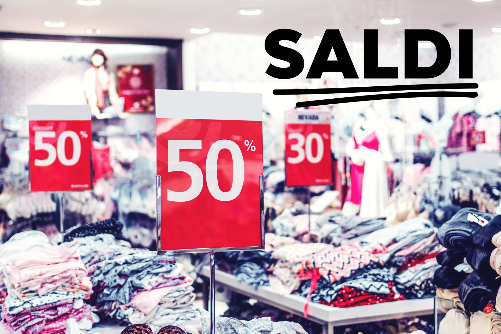 Zdjęcie przedstawia sklep odzieżowy. Po prawej stronie znajduje się napis: SALDI. Widoczne jest kilka stołów, na których ułożone są kolorowe ubrania. Na stołach znajduja się plakaty na metalowych słupkach, na których widać zapis 50 procent lub 30 procent. 