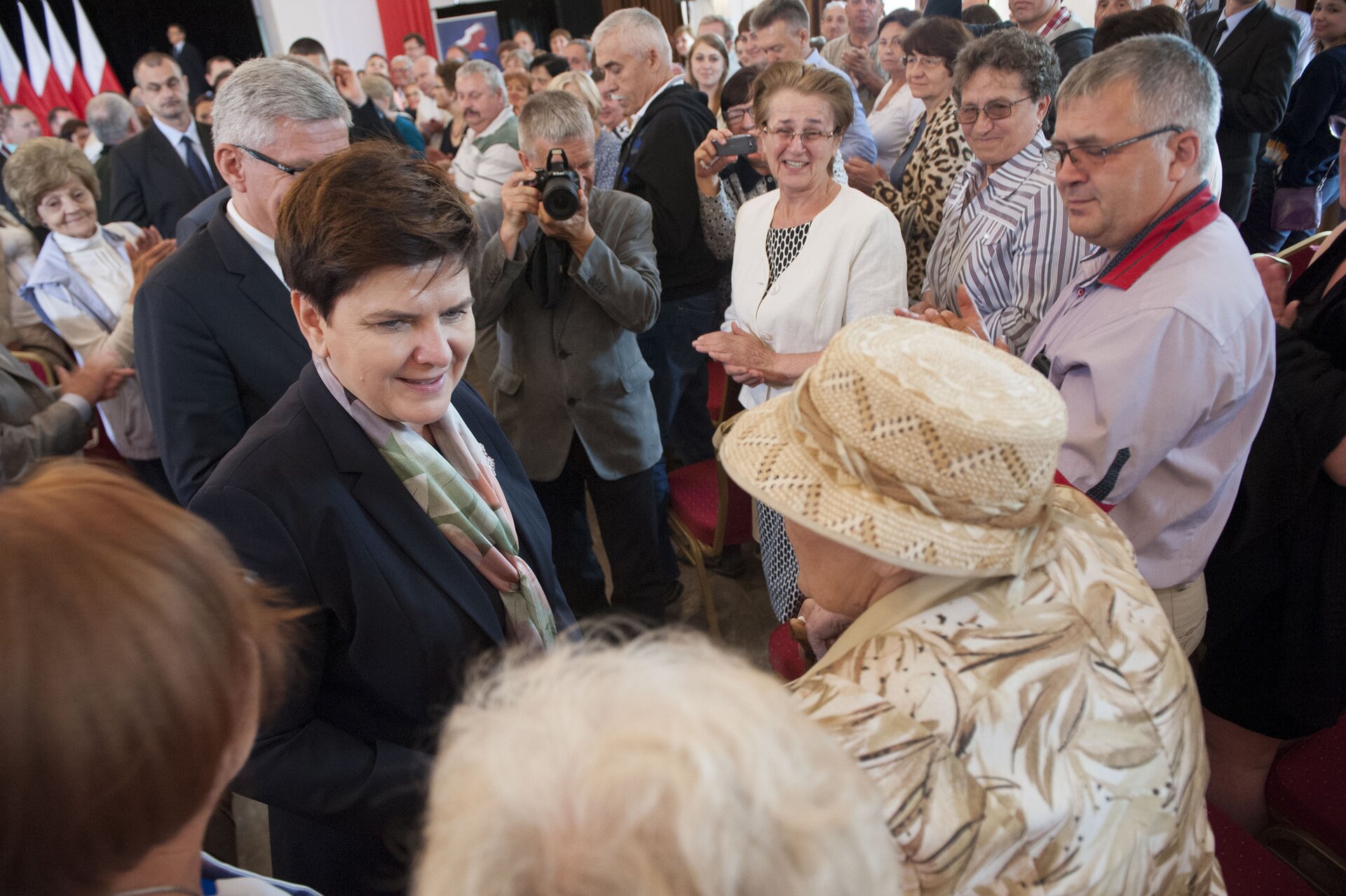 Fotografia barwna przedstawiająca spotkanie premier Beaty Szydło z repatriantami. Pani premier wita się i rozmawia ze starszymi osobami.