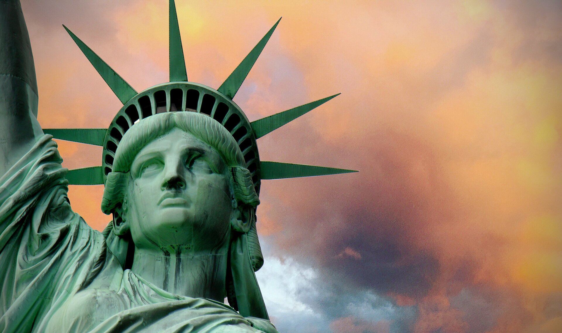 Zdjęcie ukazuje kobiecą głowę pomnika - Statuy Wolności. Na głowie jest korona z siedmioma promieniami.  W koronie znajdują się okna. Pomnika ma kolor zielonkawy. 