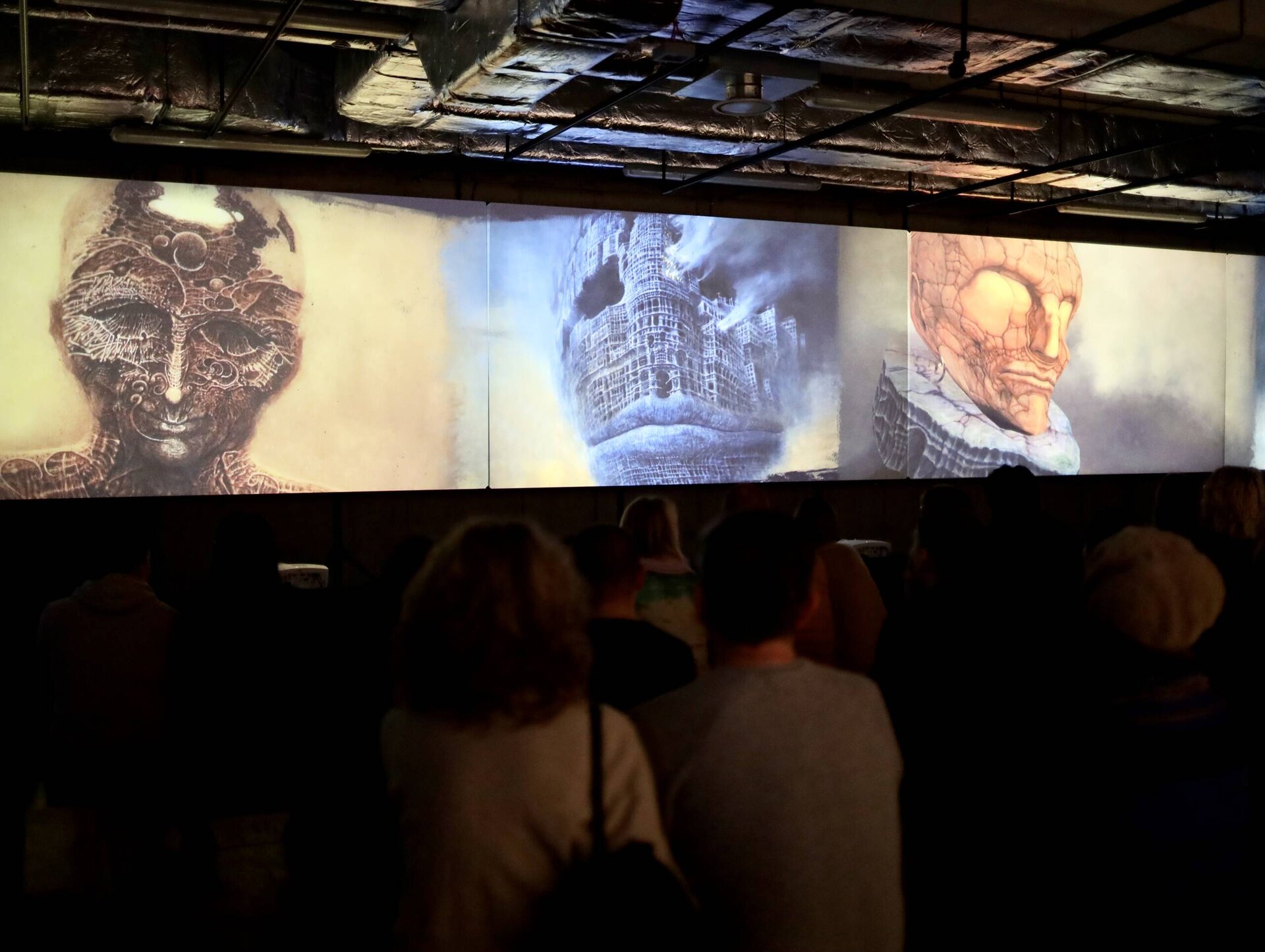 Ilustracja przedstawia kilka osób oglądających prace Zdzisława Beksińskiego na nocnej wystawie. Osoby widzą trzy dzieła artysty - wszystkie przedstawiają wizerunki osób jako surrealistyczne wizje przyszłości, a ich twarze przypominają ruiny lub konstrukcje przypominające maszyny, postacie wydają się być uwięzione w bezlitosnym świecie. Obrazy mogą wywoływać uczucia niepokoju i niepewności, wykazują się także niezwykłą wyobraźnią, techniczną precyzją i bogactwem detali. Postać na obrazie z lewej strony jest barwy brązowej, po środku – niebieskiej, a po prawej stronie pomarańczowej. Na suficie pomieszczenia, w którym miejsce ma wystawa znajdują się duże rury systemu wentylacyjnego.