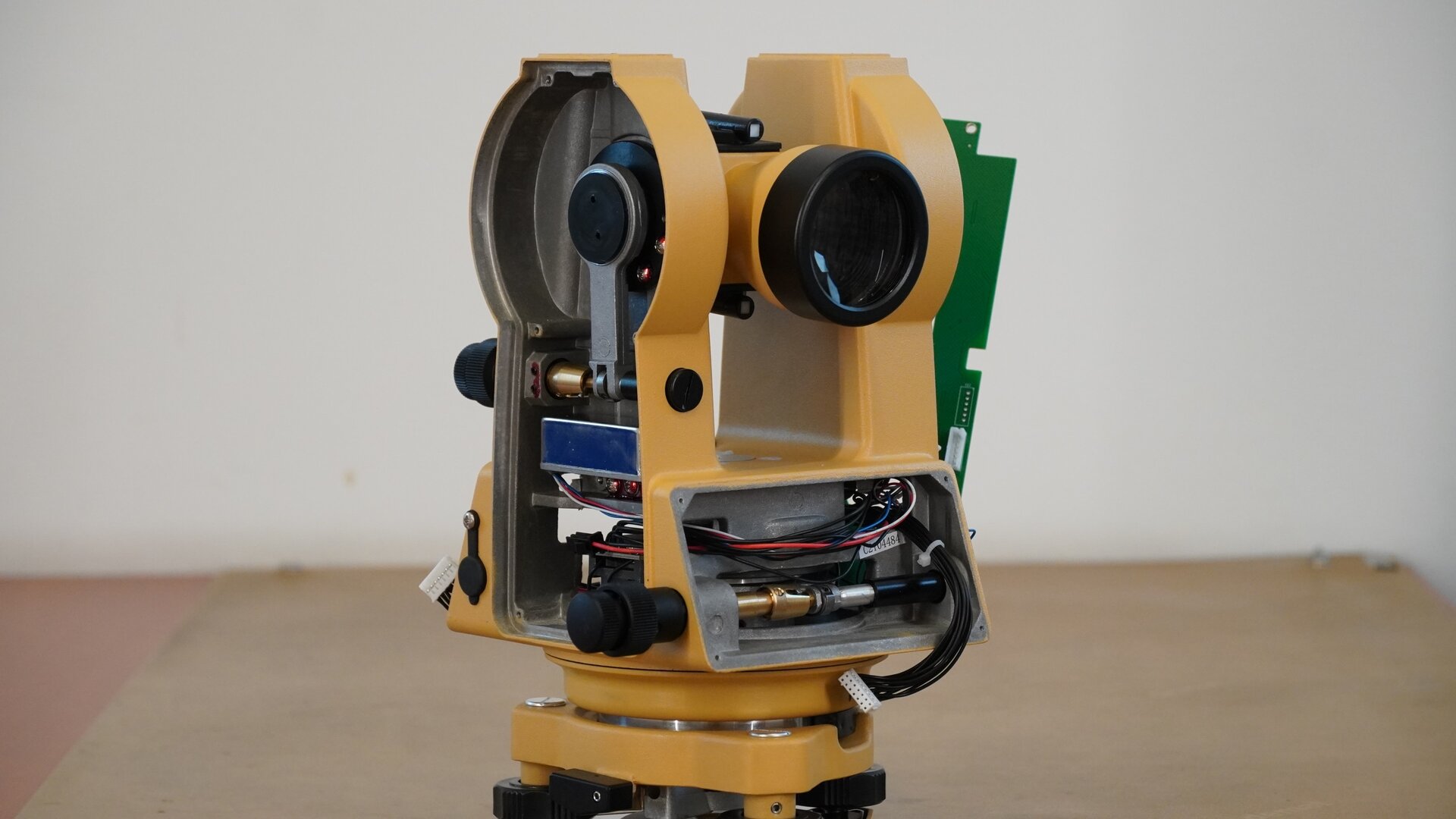 Zdjęcie ukazuje boczne ujęcie instrumentu pomiarowego od strony obiektywu. Pod obiektywem znajduje się miejsce, w którym był zamontowany wyświetlacz. Po jego demontażu w tym miejscu widoczne są cienkie przewody oraz mechanizm sterowany pokrętłem śruby leniwej. Na lewo zaś znajduje się wewnętrzny mechanizm koła pionowego z elementem zaciskowym, a poniżej kompensator elektroniczny.