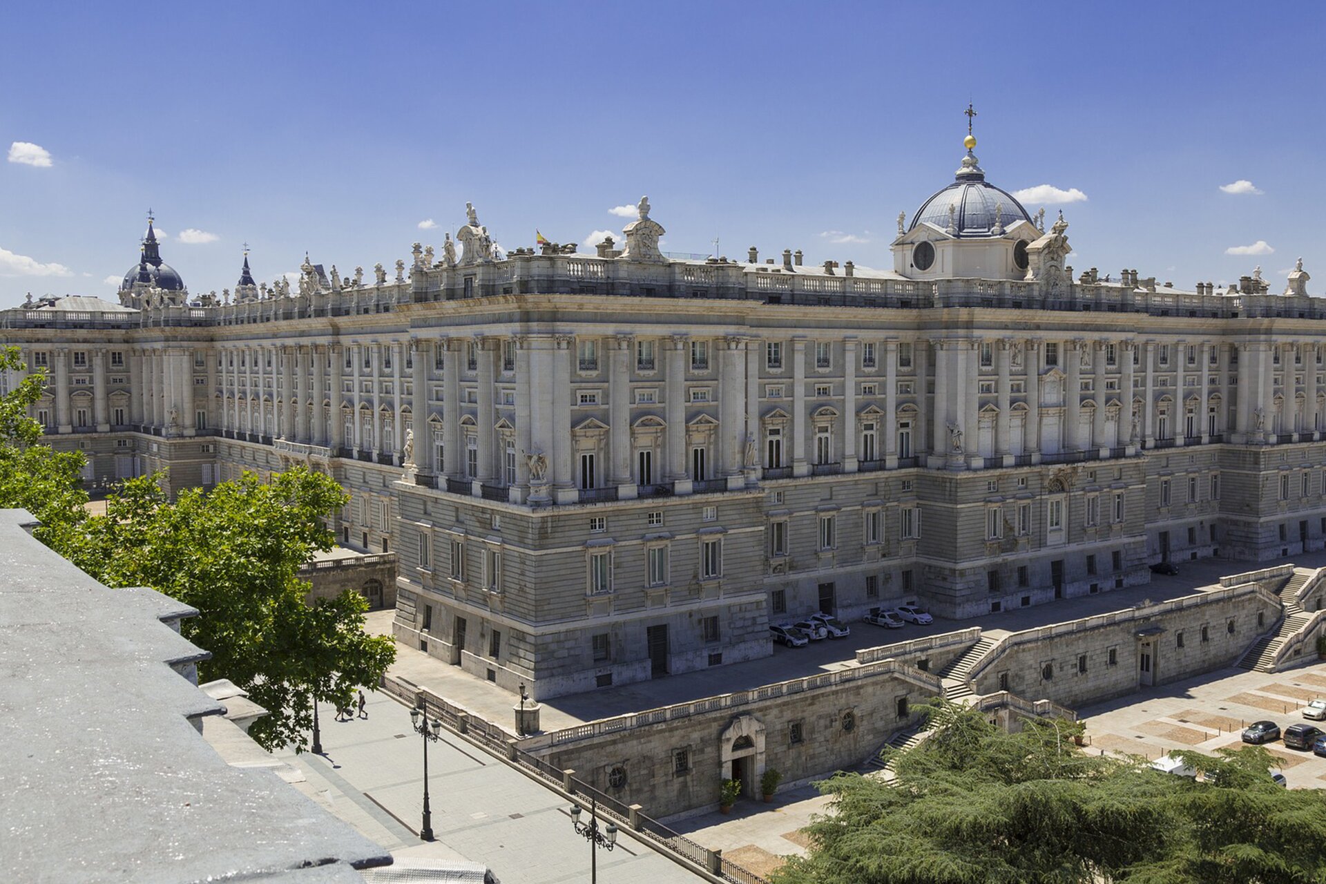 Na zdjęciu widoczny jest Palacio Real w Madrycie. Zdjęcie zrobione jest w ciepły dzień. Pałac widoczny jest na tle niebieskiego nieba. Dookoła rosną drzewa. 