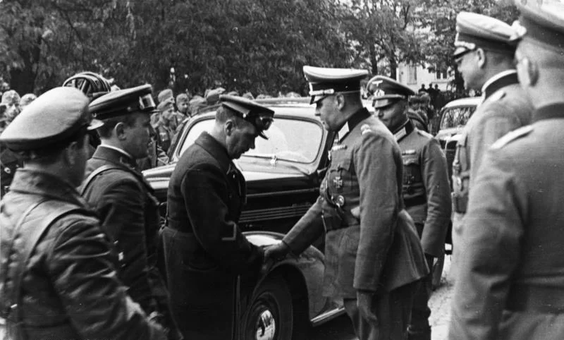 Czarno‑białe zdjęcie przedstawia dwie grupy żołnierzy, którzy stoją naprzeciwko siebie przed samochodem. Przedstawiciele każdej z grup ściskają sobie ręce. Po lewej są to żołnierze Armii Czerwonej. Mają ciemne mundury z czapkami na głowach. Po prawej są żołnierze niemieccy. Mają jaśniejsze mundury i nieco większe czapki. W tle stoją w szeregu żołnierze.