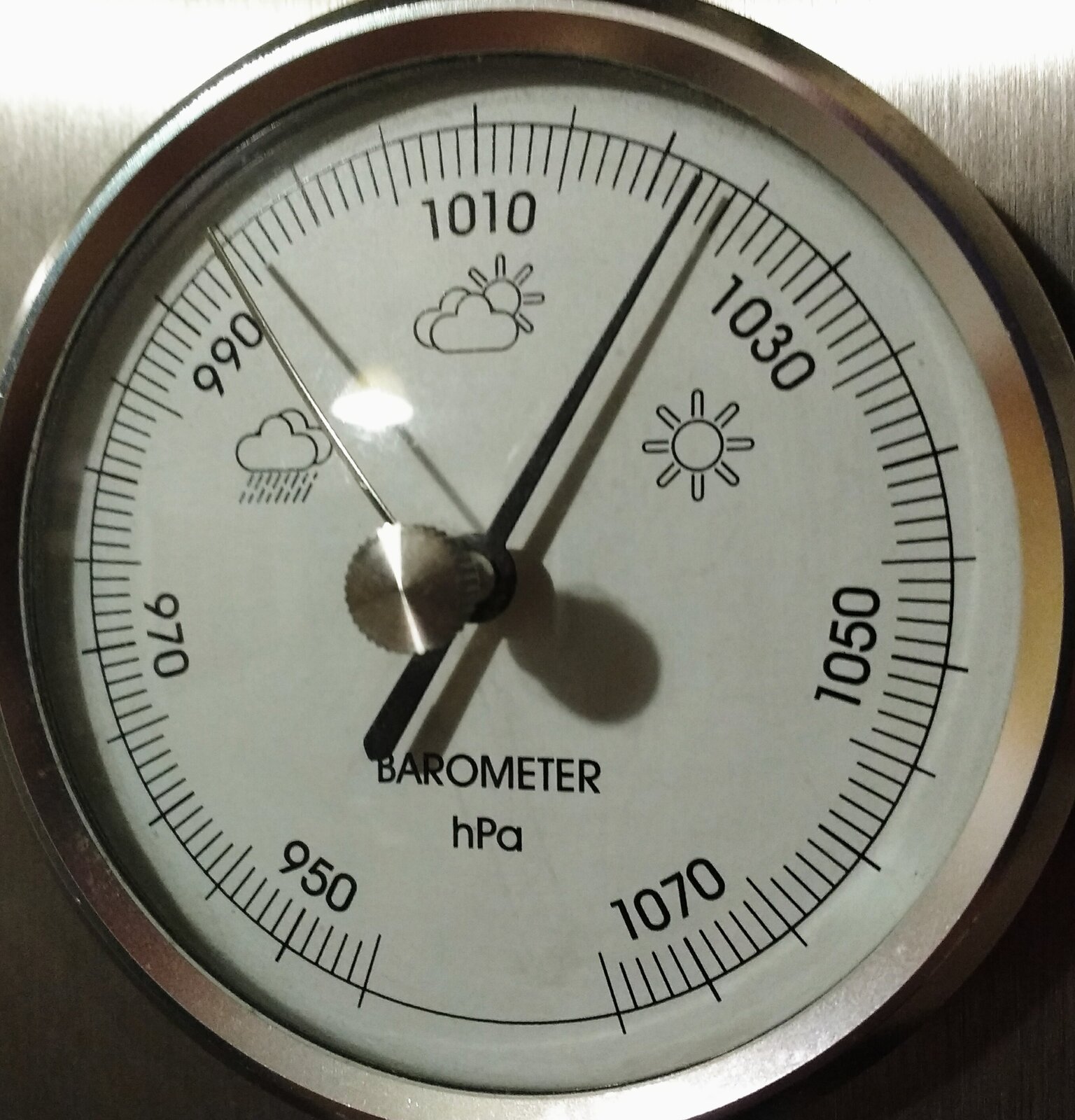 Zdjęcie przedstawia barometr mechaniczny. Jest to przyrząd o okrągłej tarczy. Na tarczy widać podziałkę z wartościami podanymi w hektopaskalach. Wskazówka barometru ukazuje wartość ciśnienia powietrza.