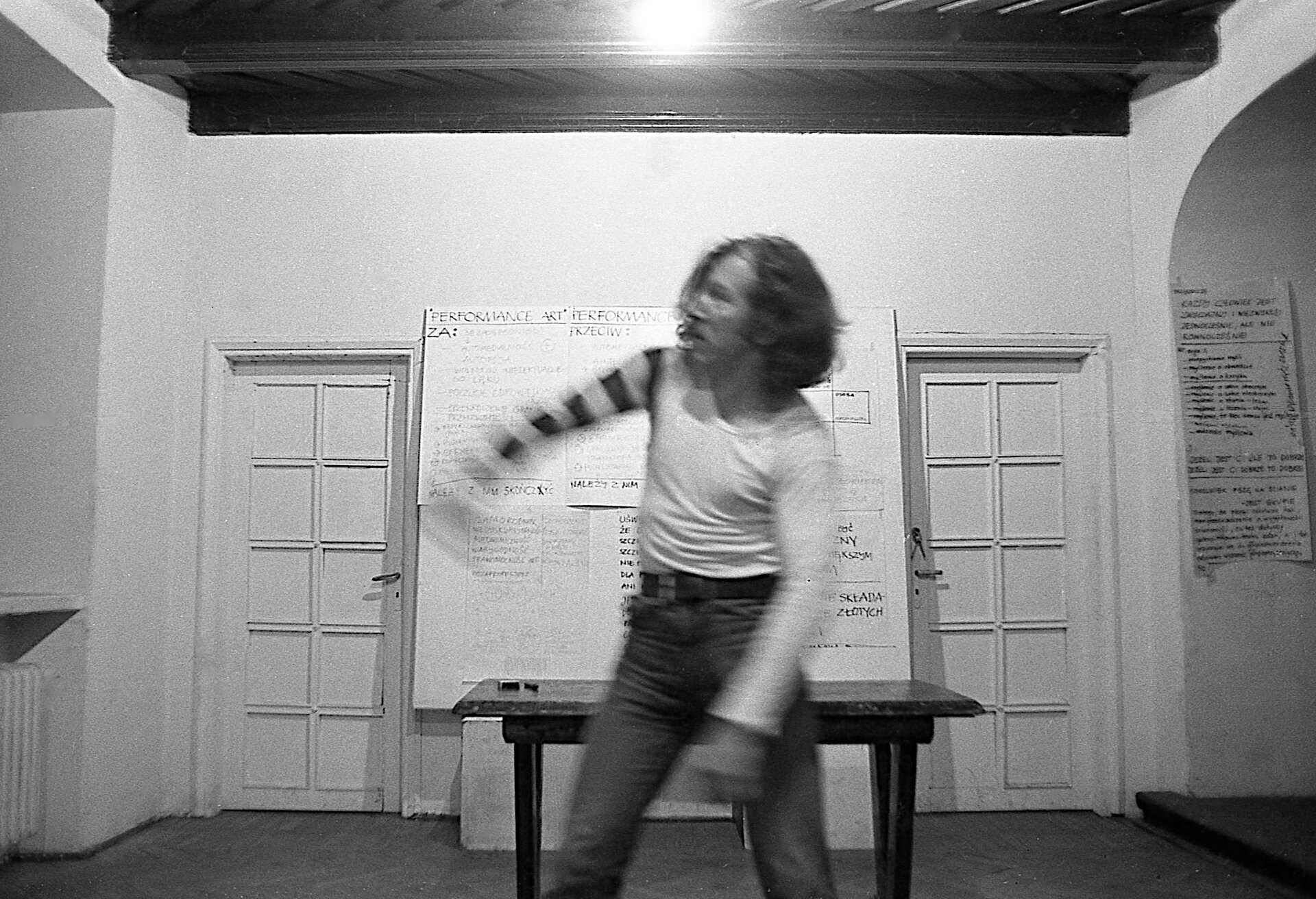 Ilustracja w formie prostokąta przedstawia czarno‑białą fotografię. Na pierwszym planie ukazany został mężczyzna o włosach średniej długości. Ubrany jest w białą koszulkę na długi rękaw, której jeden z rękawów posiada czarne paski praz ciemne spodnie. Wzrok oraz głowa mężczyzny skierowane są w lewą stronę. Jedną z rąk unosi delikatnie ku górze. Jest ona rozmazana na fotografii. W tle ukazuje się jasna ściana, na której umiejscowione są dwie pary jasnych drzwi. Pomiędzy nimi zawieszona jest duża, biała plansza z czarnymi napisami. Przed nią ustawiony jest ciemny stół. Przy suficie znajdują się ciemne belki. 