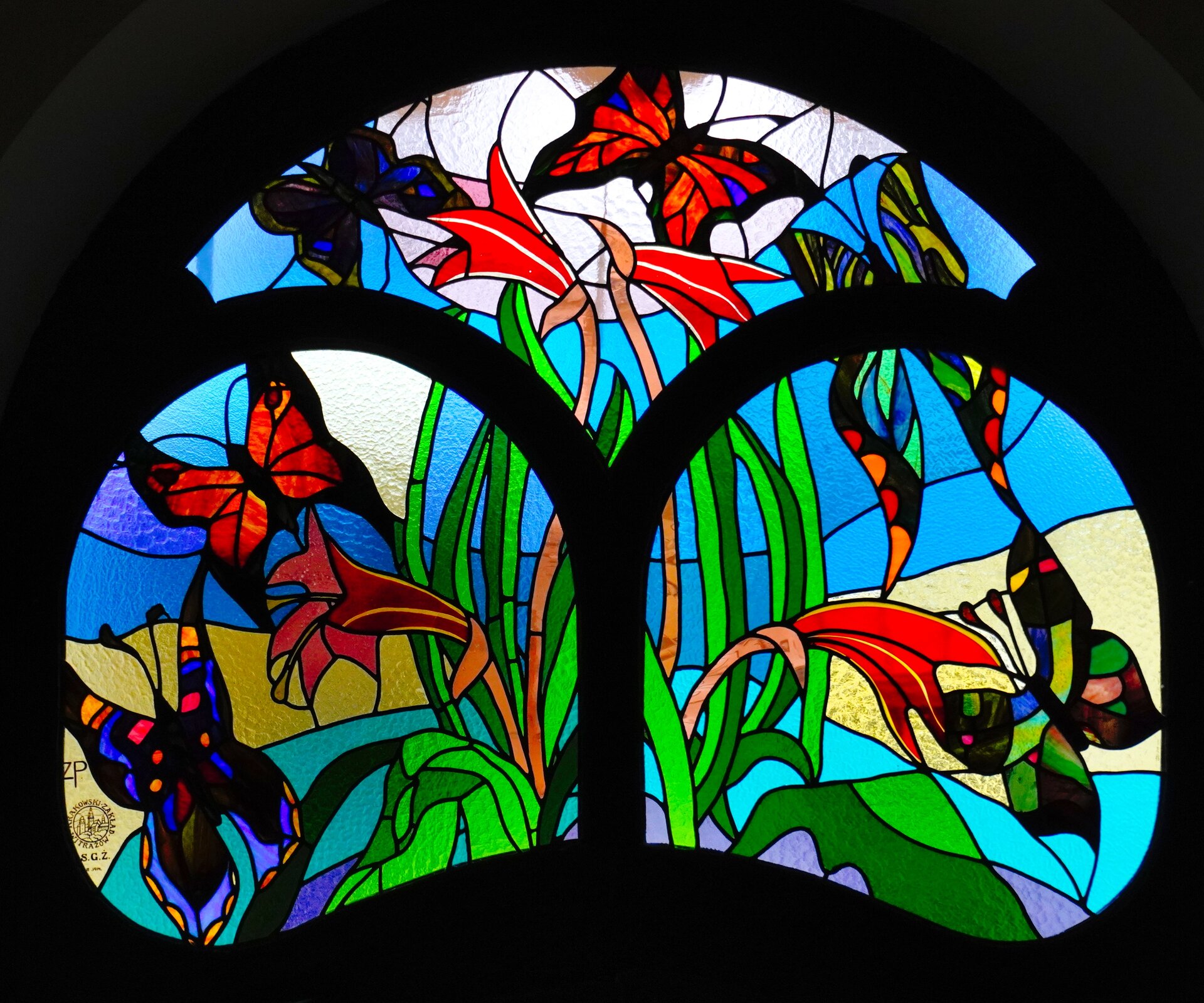 Witraż jest wykonany z kolorowych szkiełek i przedstawia motyle latające wokół kwiatów. Każde szkiełko obramowane jest czarnym, ołowianym konturem. Witraż podzielony jest na trzy części. Z lewej strony znajduje się motyl o kolorach: niebieskim, fioletowym, pomarańczowym i czerwonym. Nad nim - motyl czerwony. W górnej części znajduje się motyl w ciemnych kolorach, a obok niego - motyl czerwony z dwoma niebieskimi akcentami. Pod nim lecą jeszcze dwa motyle. Rośliny mają długie łodygi w jasnozielonym i ciemnozielonym kolorze i czerwone, podłużne kwiaty w kształcie dzwonków. Tło jest utworzone z różnokolorowych szkiełek - niebieskich, fioletowych, białych.