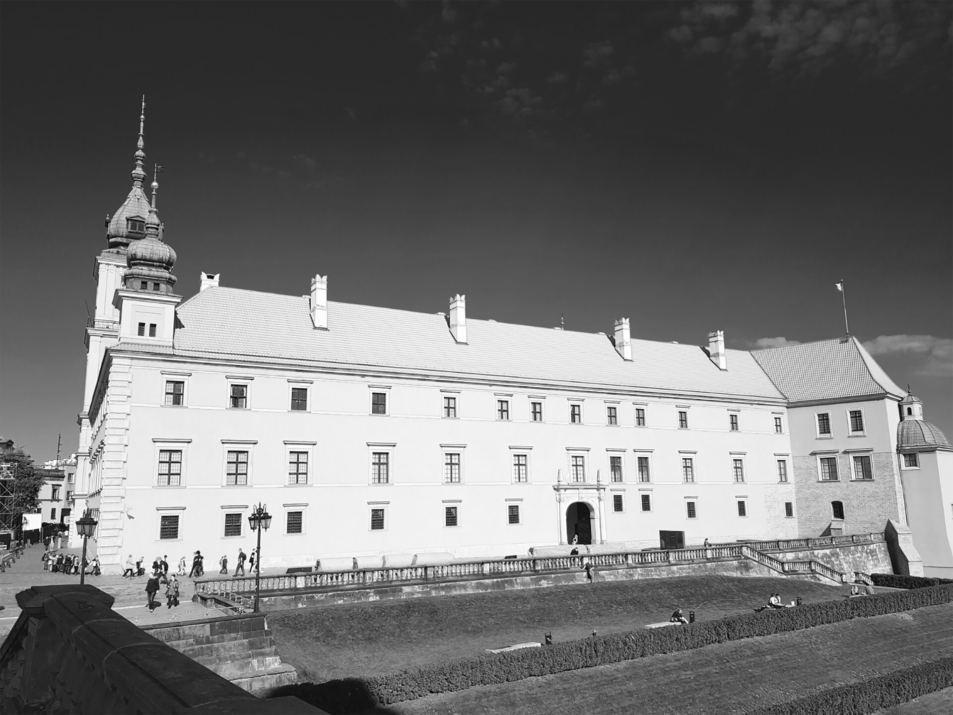 Zdjęcie czarno‑białe przedstawiający zamek, którego ściany są białe, szarym oznaczone są okna i ich krawędzie, dach ciemniejszy jest od ścian. Niemal całkowicie czarne niebo oraz ciemnoszara trawa.