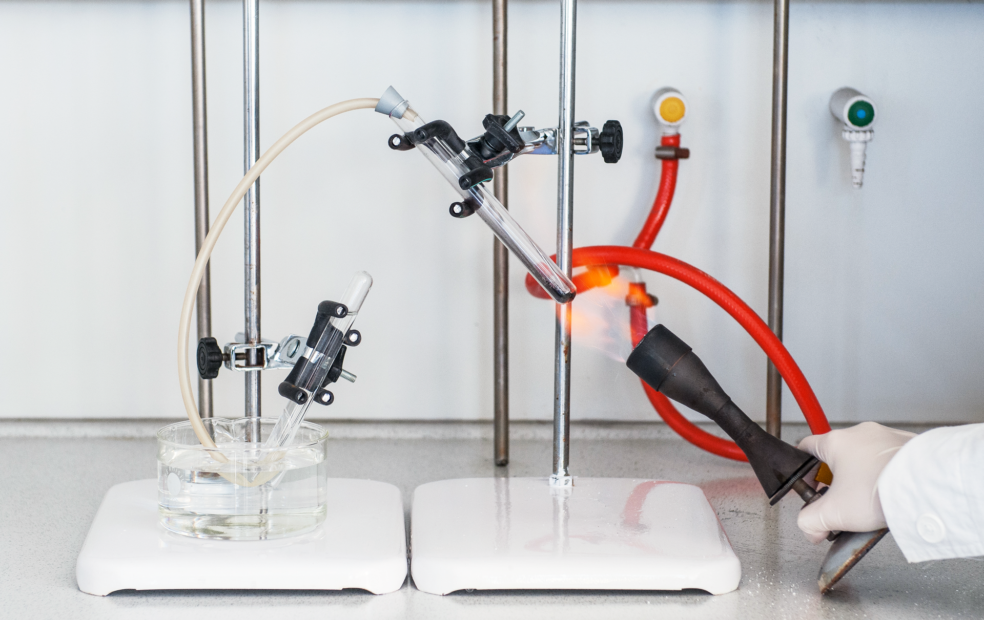 Ilustracja przedstawia zdjęcie aparatury do otrzymywania tlenu w warunkach laboratoryjnych. Składa się ona z dwóch stojących obok siebie statywów z łapami w których zamocowano probówki. Statyw z prawej strony trzyma długą probówkę ze znajdującymi się w niej fioletowymi drobnymi kryształkami otworem w górę pod kątem około 45 stopni do pionu. Otwór jest zakorkowany, a przez korek przeciągnięta jest gumowa rurka prowadząca do aparatury z lewej strony. Probówka na statywie z lewej strony jest obrócona otworem w dół pod kątem około 30 stopni do pionu. Koniec probówki zanurzony jest w szerokiej szklanej zlewce z wodą. Sama probówka również wypełniona jest wodą. Koniec wężyka z probówki po prawej stronie umieszczony jest w odwróconej probówce po lewej stronie. Dłoń demonstratora trzyma palnik gazowy w taki sposób, że płomień ogrzewa dno probówki po lewej i znajdujące się wewnątrz kryształki. Wydzielający się gaz przepychany przez rurkę zbiera się w probówce po lewej stronie wypierając z niej wodę.