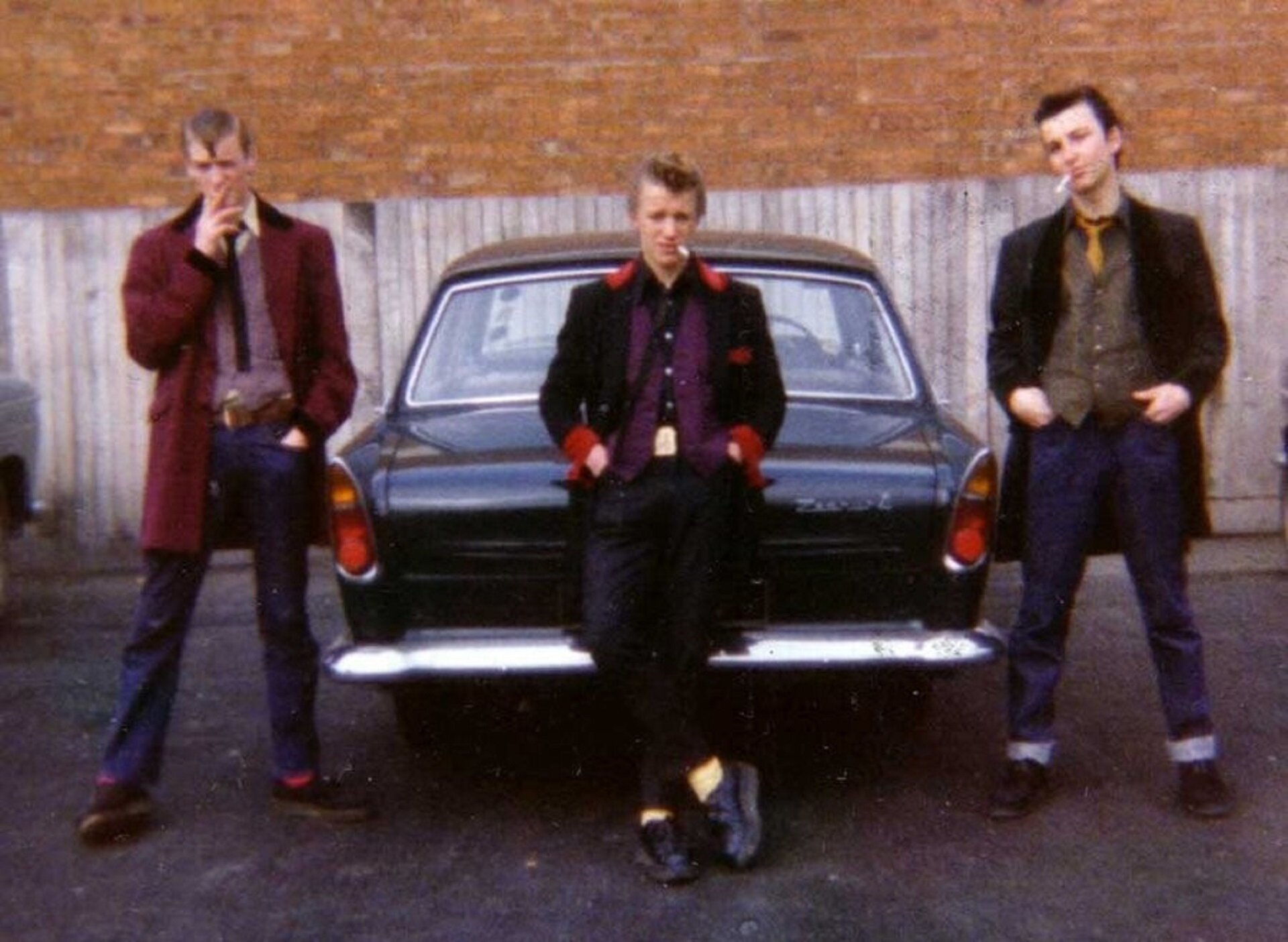 Ilustracja przedstawia trzech młodych mężczyzn na tle czarnego samochodu. Palą papierosy i mają ręce w kieszeniach. Mężczyzna w środku opiera się o bagażnik, ma na sobie żółto-czarny krawat, żółte skarpetki, czarne spodnie i buty, czarną, długą marynarkę z czerwonymi mankietami i kołnierzem oraz fioletową koszulę. Mężczyzna stojący po lewej stronie ma na sobie białą koszulę, fioletowy sweter, bordowe skarpetki i długą, bordową marynarkę z czarnymi mankietami i kołnierzem, czarny krawat i buty oraz niebieskie, dżinsowe spodnie. Mężczyzna stojący po prawej stronie ma na sobie żółty krawat, brązową kamizelkę, czarną długą marynarkę i buty oraz niebieskie, dżinsowe spodnie.