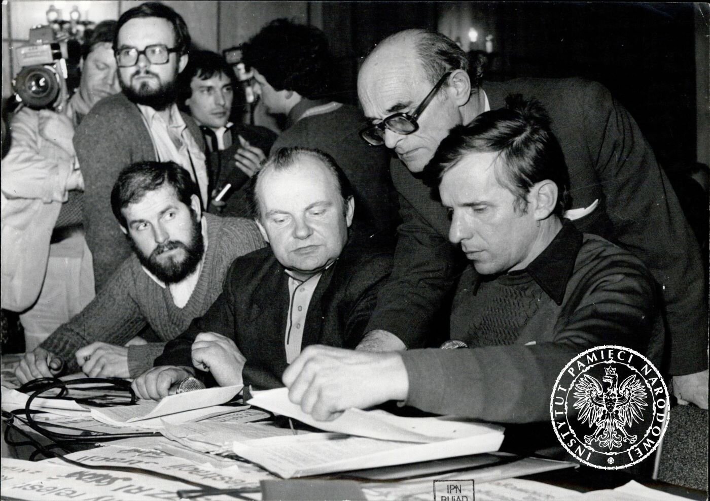 Zdjęcie przedstawia mężczyzn skupionych i na dokumentach leżących na stole przed nimi. Czytają je, studiują. Za nimi stoją dziennikarze z kamerą, aparatem, mikrofonem.
