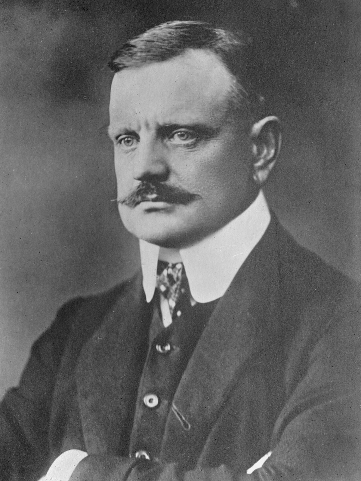 Ilustracja przedstawia fotoportret Jeana Sibeliusa autorstwa Daniela Nyblina. Zdjęcie pochodzi z 1913 roku. Mężczyzna siedzi ze skrzyżowanymi rękoma na wysokości klatki piersiowej. Minę ma poważną, lekko zmarszczone okolice między brwiami. 