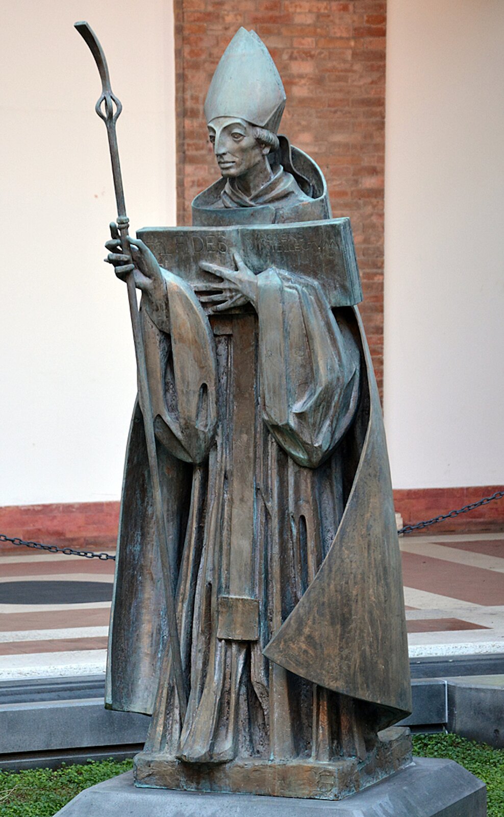 Zdjęcie przedstawia pomnik mężczyzny w długiej szacie duchownego. Ma na głowie mitrę biskupią, w rękach trzyma pastorał oraz księgę.