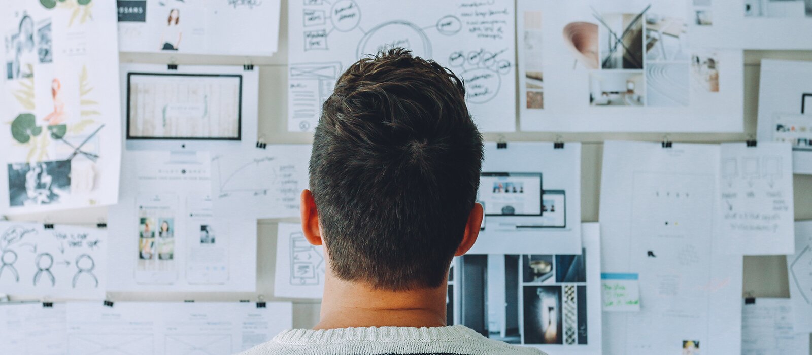 Zdjęcie przedstawia mężczyznę, w ujęciu od tyłu, od ramion w górę, który stoi przed ścianą, na której są poprzypinane różne kartki z informacjami, rysunkami i zdjęciami. 